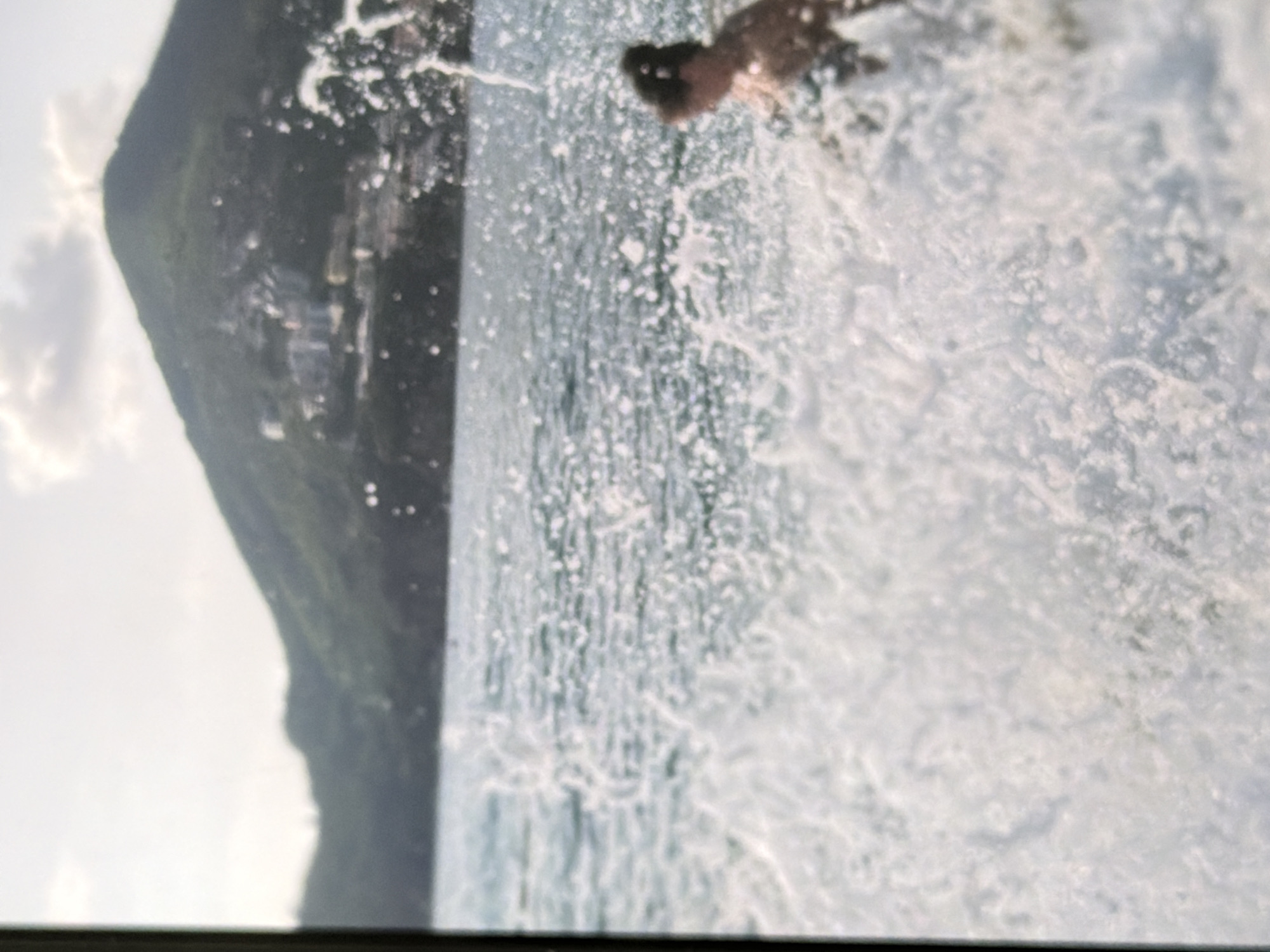 A person is partially visible in the right side of the frame, in profile and facing left, with their upper body submerged in water. They appear to be in the ocean or a large body of water, as evidenced by the waves and splashes surrounding them. In the background, a lush green mountain rises from the water's edge, with buildings scattered across its slopes. The sky above is bright with some clouds. The image is slightly out of focus, contributing to a sense of motion and dynamism.