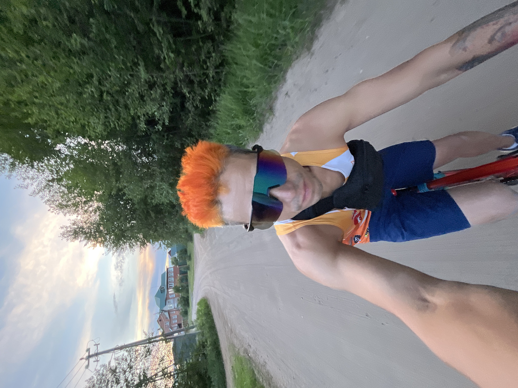 This is a first-person perspective photograph taken by a person riding a bicycle on a dirt road. The rider has bright orange hair and is wearing athletic sunglasses with a mirrored, iridescent lens, a yellow and white tank top, and blue cycling shorts. A black fanny pack is worn around their waist. The road is flanked by green trees and grass. In the background, several houses with red roofs are visible, suggesting a rural or suburban setting. The sky is partly cloudy with a soft, warm light, indicating it might be late afternoon or early evening. The overall mood appears casual and outdoorsy. There is no visible text in the image.