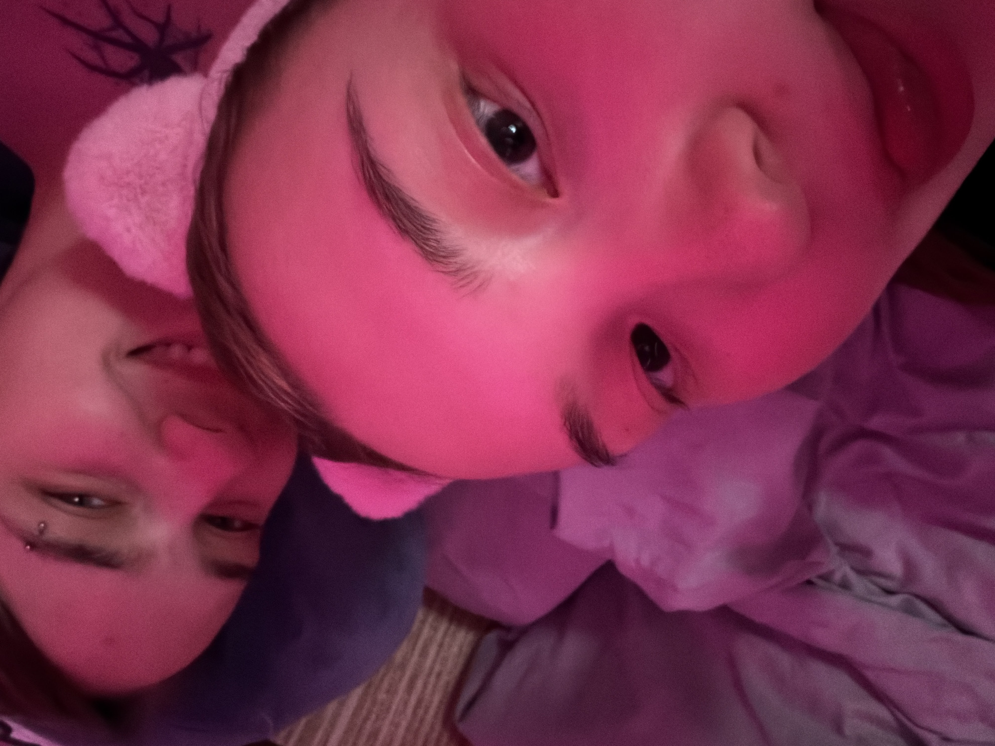 Two individuals are visible in the image, illuminated by a strong pink light source, likely artificial. One individual, positioned in the upper right quadrant of the image, is wearing a headband with fuzzy, possibly animal-themed, ears. Their face is turned towards the camera, revealing one eye, an eyebrow, part of their nose, and a portion of their mouth.
The second individual is located on the left side of the frame. They are also facing the camera and are partially smiling. A possible eyebrow piercing is visible above one eye.
The background consists of various textures, including a pillow and a draped fabric, suggesting an indoor setting. The setting is likely a domestic environment. The image was supposedly taken in Chaikovsky, Russia, though there is no visual evidence to confirm this.