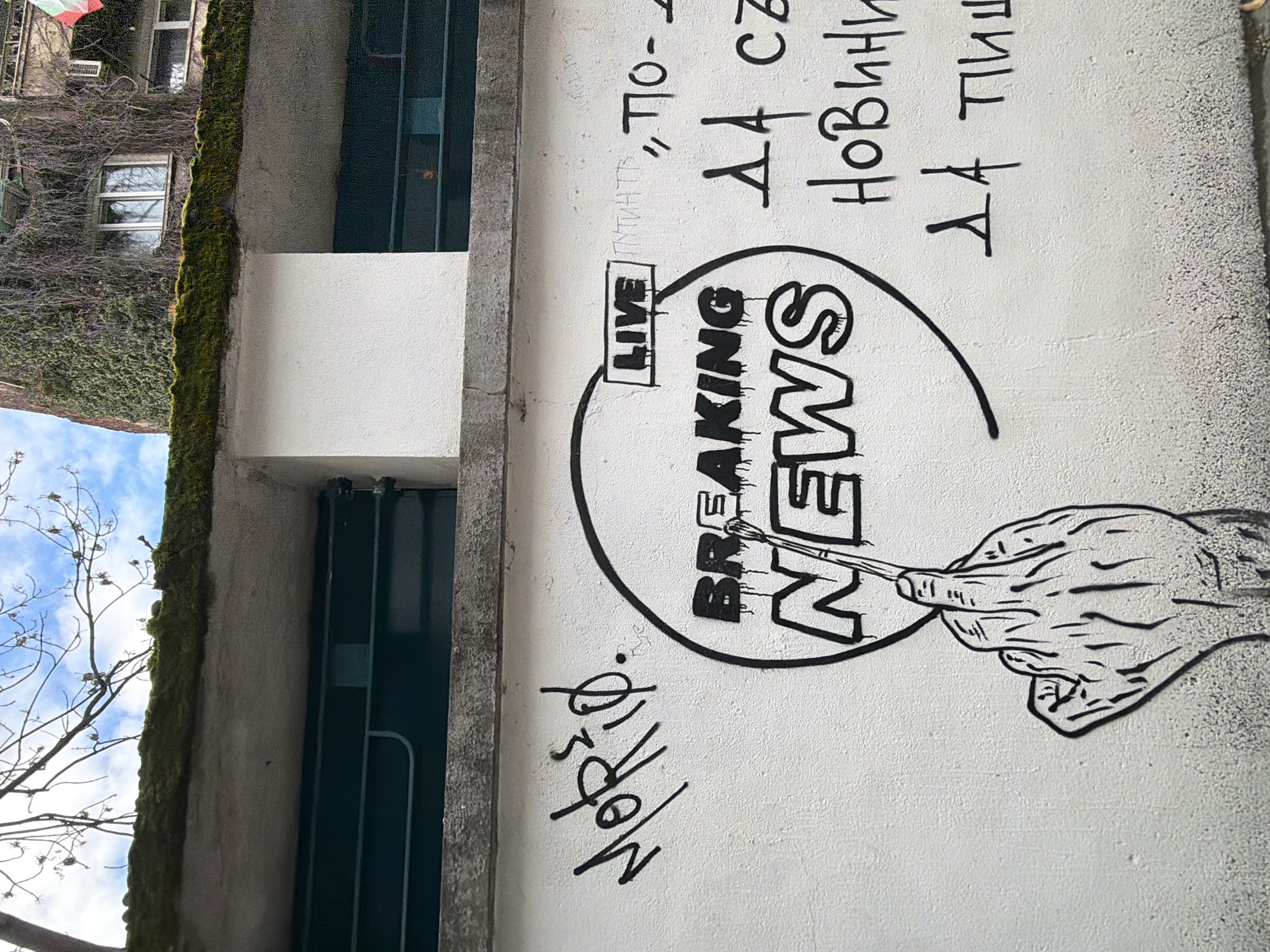 The image shows street art on a white wall in Sofia, Bulgaria. The wall appears to be part of a building exterior, with green moss growth visible along the top edge. The art features the words "BREAKING NEWS" inside a circle, with the word "LIVE" in a box above it. A hand holding a paintbrush is also depicted, seemingly painting the "N" in "NEWS." Above the artwork, there are handwritten phrases in Bulgarian and other graffiti. To the left, there is a closed green security gate. The sky is visible with some tree branches in the lower left corner.
