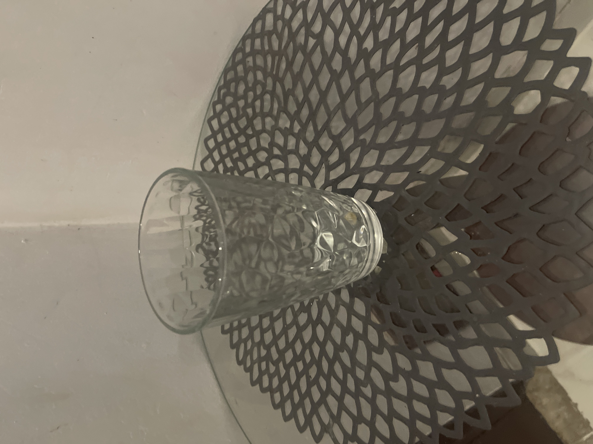 The image shows a glass tumbler resting on a round, grey placemat with a decorative, cutout pattern. The placemat is positioned on a flat surface, possibly a table, with a white wall in the background. The tumbler itself is made of clear glass and has a textured surface, appearing to have a diamond-like pattern etched into it. The light source, which is not visible in the frame, casts a distinct shadow of the placemat onto the wall behind it. The shadow mimics the intricate cutout design of the placemat, creating a visually interesting effect. The overall lighting suggests an indoor setting, possibly during the day given the natural shadows. There is no visible text in the image. The composition focuses on the interplay of light, shadow, and texture between the glass tumbler and the placemat.
