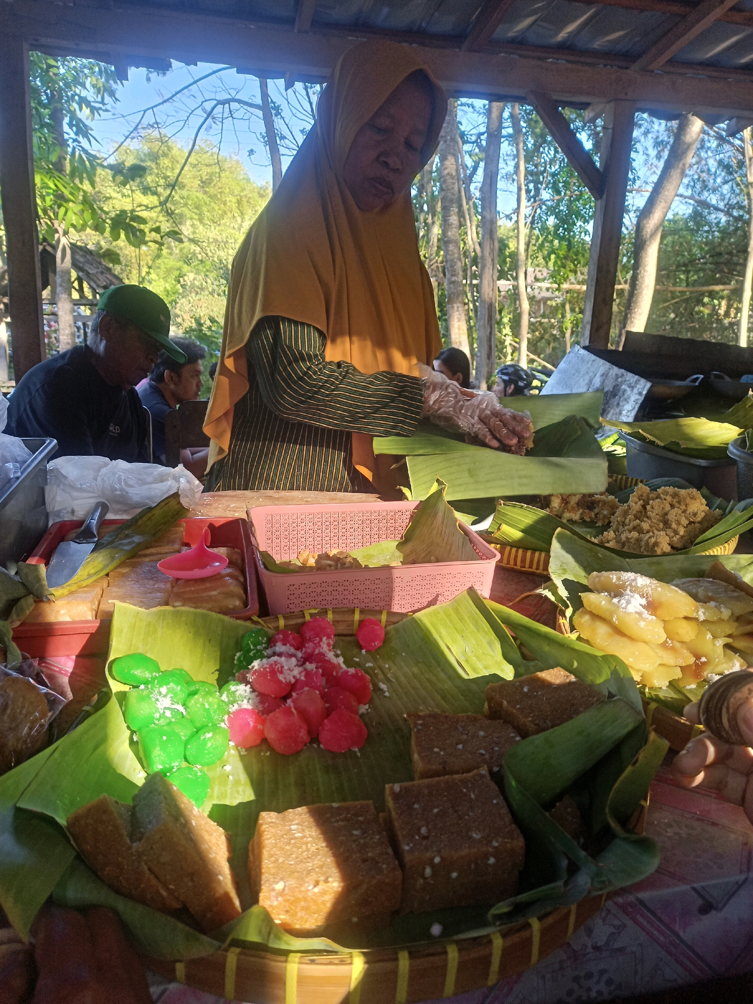 The image captures an outdoor market scene in Karungan, Indonesia, likely during daytime given the sunlight. A woman wearing a yellow hijab and striped shirt is in the background, using plastic gloves to handle food wrapped in banana leaves. In the foreground, various types of traditional Indonesian snacks are displayed on banana leaves within woven baskets and plastic containers. These include rectangular brown cakes, colorful spherical candies (pink and green), and yellow slices dusted with white powder. Several men are visible in the background, one wearing a green cap and black shirt, possibly customers. The setting appears to be a food stall under a simple wooden and corrugated metal roof, with trees and foliage visible in the background, suggesting a rural or semi-rural environment.