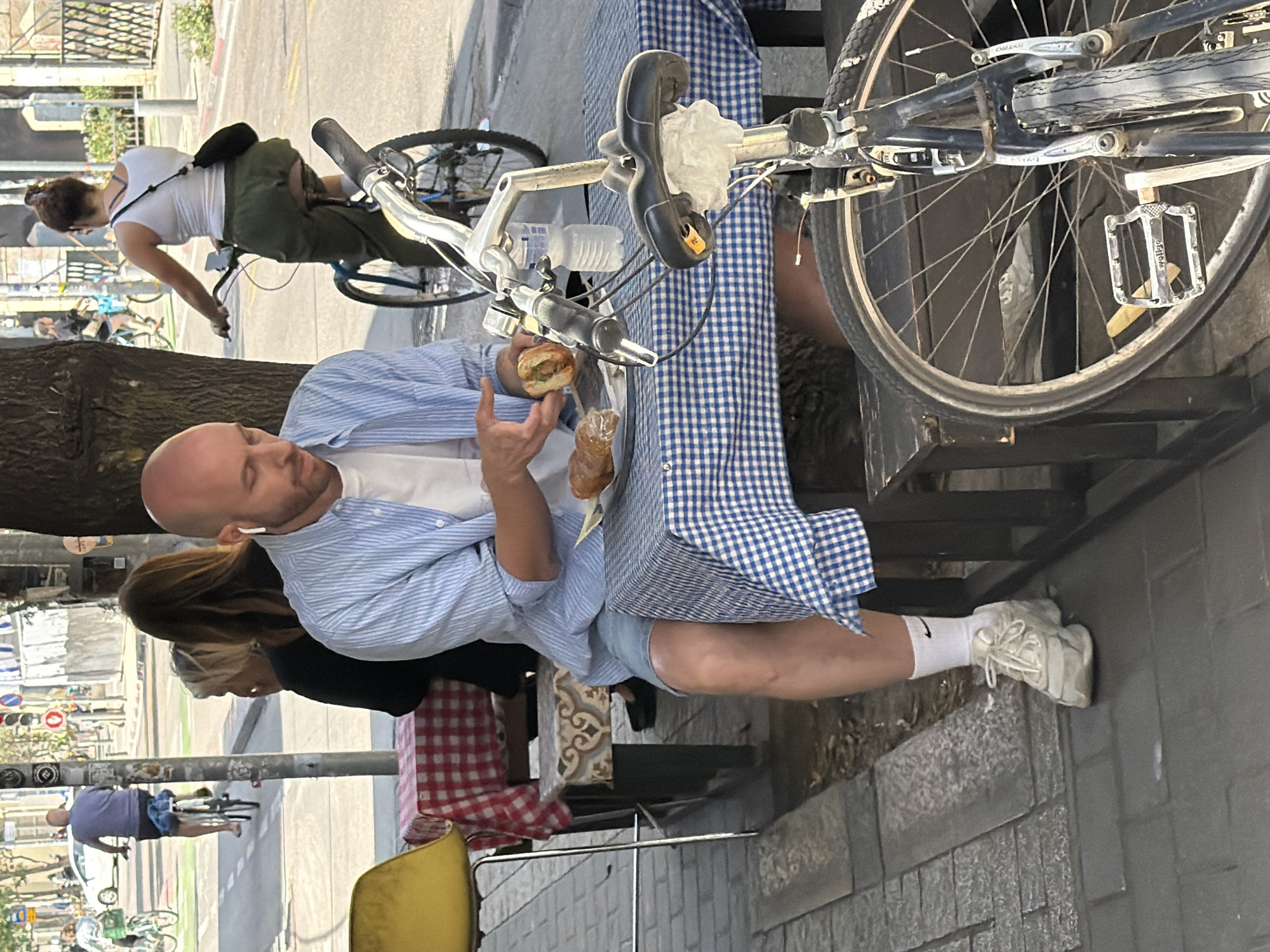 A medium shot captures a man in Tel Aviv, Israel, eating on a sunny day. He is bald with a light beard, wearing a light blue striped shirt over a white t-shirt and blue shorts, and white sneakers with white socks. He is seated at a table covered with a blue-and-white checkered cloth, holding a sandwich in one hand and a pastry in the other. A bicycle, with its front wheel prominently displayed, is parked against the table. Another woman is seated near him, wearing a black top. In the background, a woman rides a bicycle, wearing a white top and green pants, with a black bag. A tree is partially visible to the left. The location appears to be a street scene with parked bicycles.
