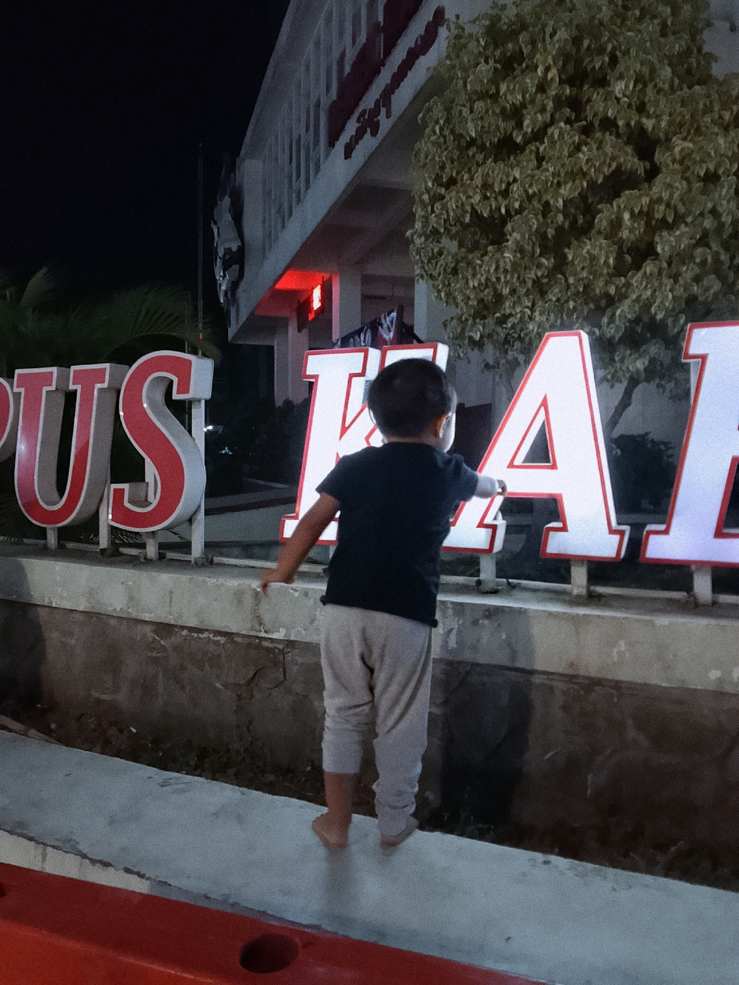 A young child wearing a dark t-shirt and light-colored pants stands with their back to the camera, reaching out to touch large, illuminated letters that spell out "PUS". The letters are red and white, with a bright red outline, and are mounted on poles. Behind the child and the letters, a building with a white facade and red accents is visible. A dense, leafy green tree is situated to the right of the child, partially obscuring the building. The scene is illuminated by artificial lights, suggesting it is nighttime. A red barrier or structure is visible in the foreground. The location is identified as Boyolali, Indonesia.