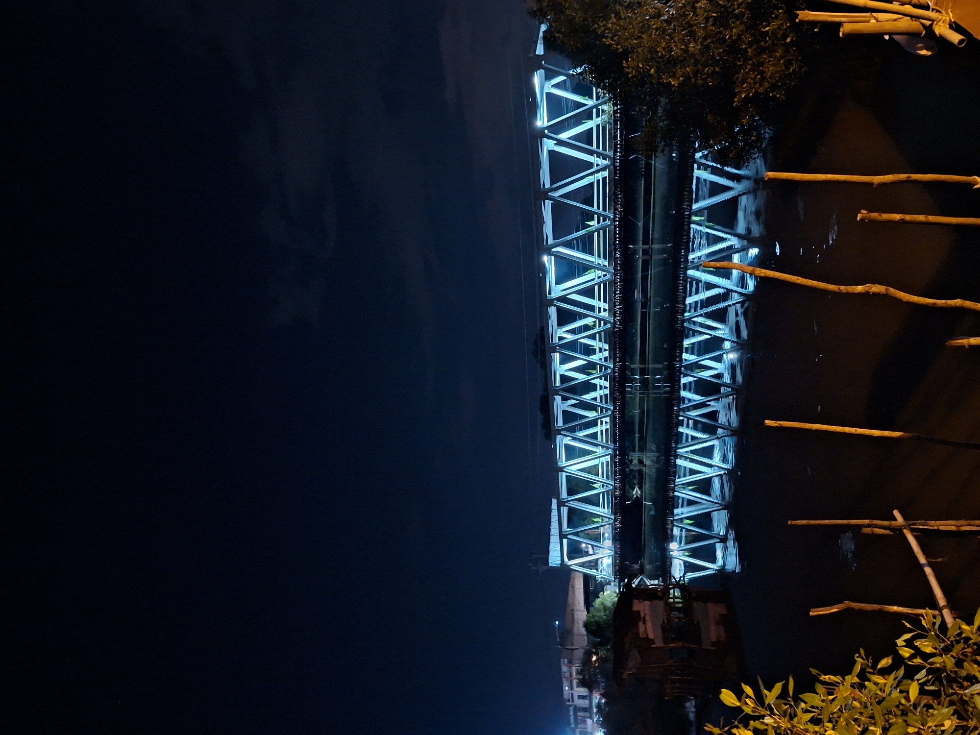 The image shows a bridge at night in Lhokseumawe, Indonesia. The bridge is brightly lit with blue lights outlining its triangular support structure. The sky is dark and largely empty, occupying the left side of the frame. Below the bridge is water that appears dark, reflecting some of the blue light. Along the water's edge are wooden posts and some vegetation. Buildings and lights from the city can be seen faintly in the bottom left. The image seems to be taken from a low angle, looking up at the bridge.