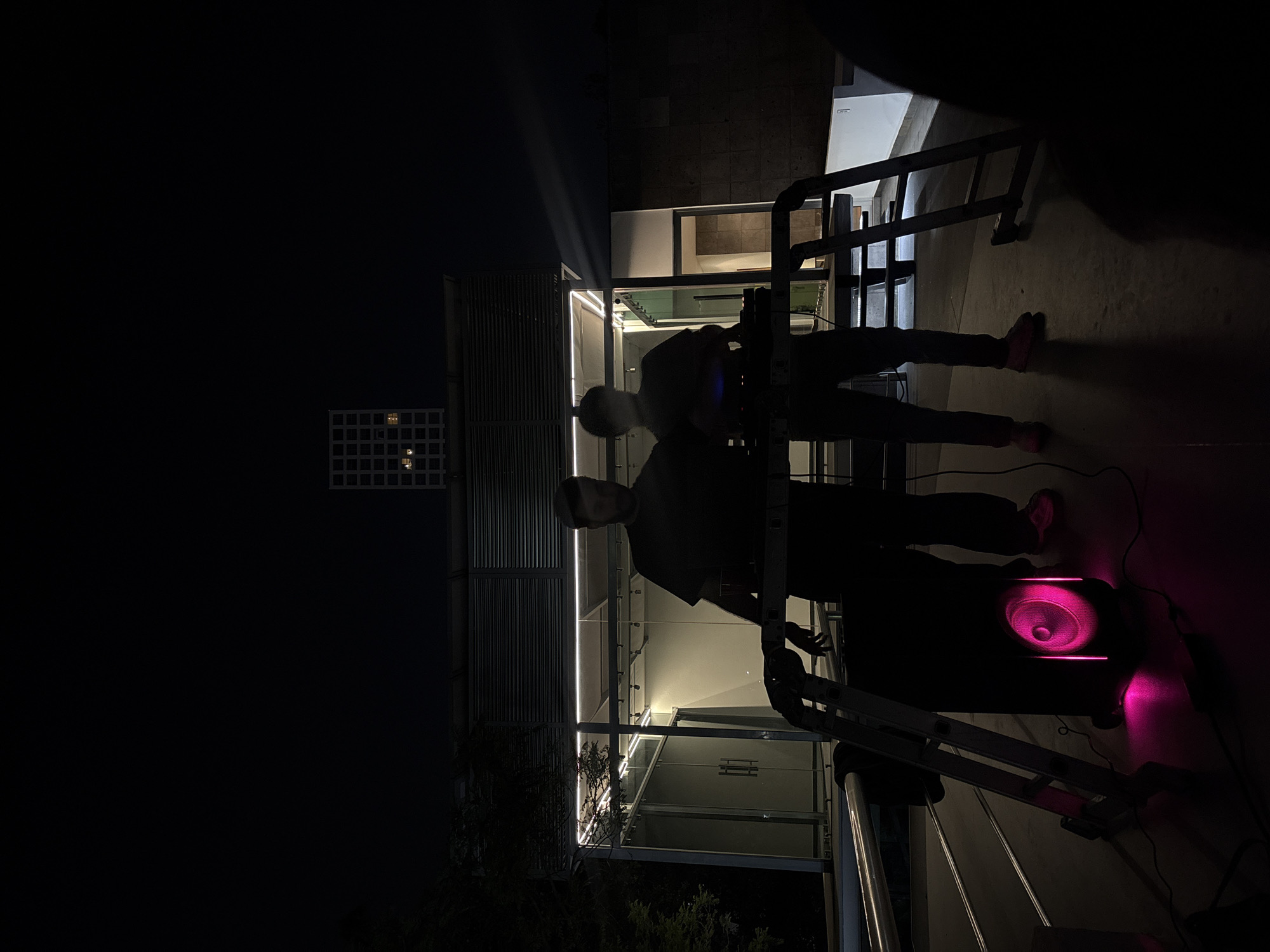 The image, taken at night, features a man standing on a ladder, seemingly operating sound equipment. The location appears to be outdoors near a modern building with glass panels and linear lighting. A speaker is visible, illuminated with a pink light. The background is dark, but a building with a latticework design is visible in the distance. The presence of sound equipment suggests the setup for an event or performance. It's impossible to confirm the exact location in San Bernardino Tlaxcalancingo without more specific details.