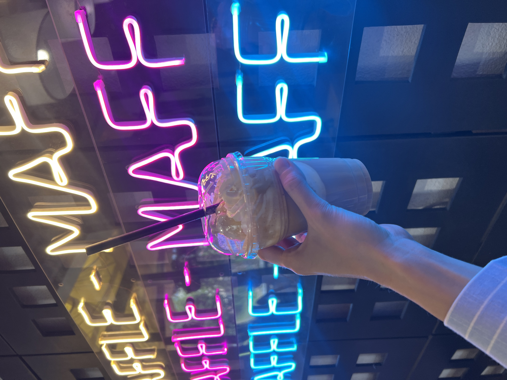 A hand is holding a disposable cup with a straw in front of a mirrored surface. Reflected in the mirror are neon signs that spell out "CAFE". The neon signs are lit in yellow, pink, and blue. The background consists of a grid-like pattern of small, dark rectangles. The scene appears to be taken indoors, possibly at a cafe or bar in Bar, Montenegro, given the "CAFE" sign in English. The cup appears to contain a beverage with whipped cream. The hand is wearing a light-colored, striped sleeve.