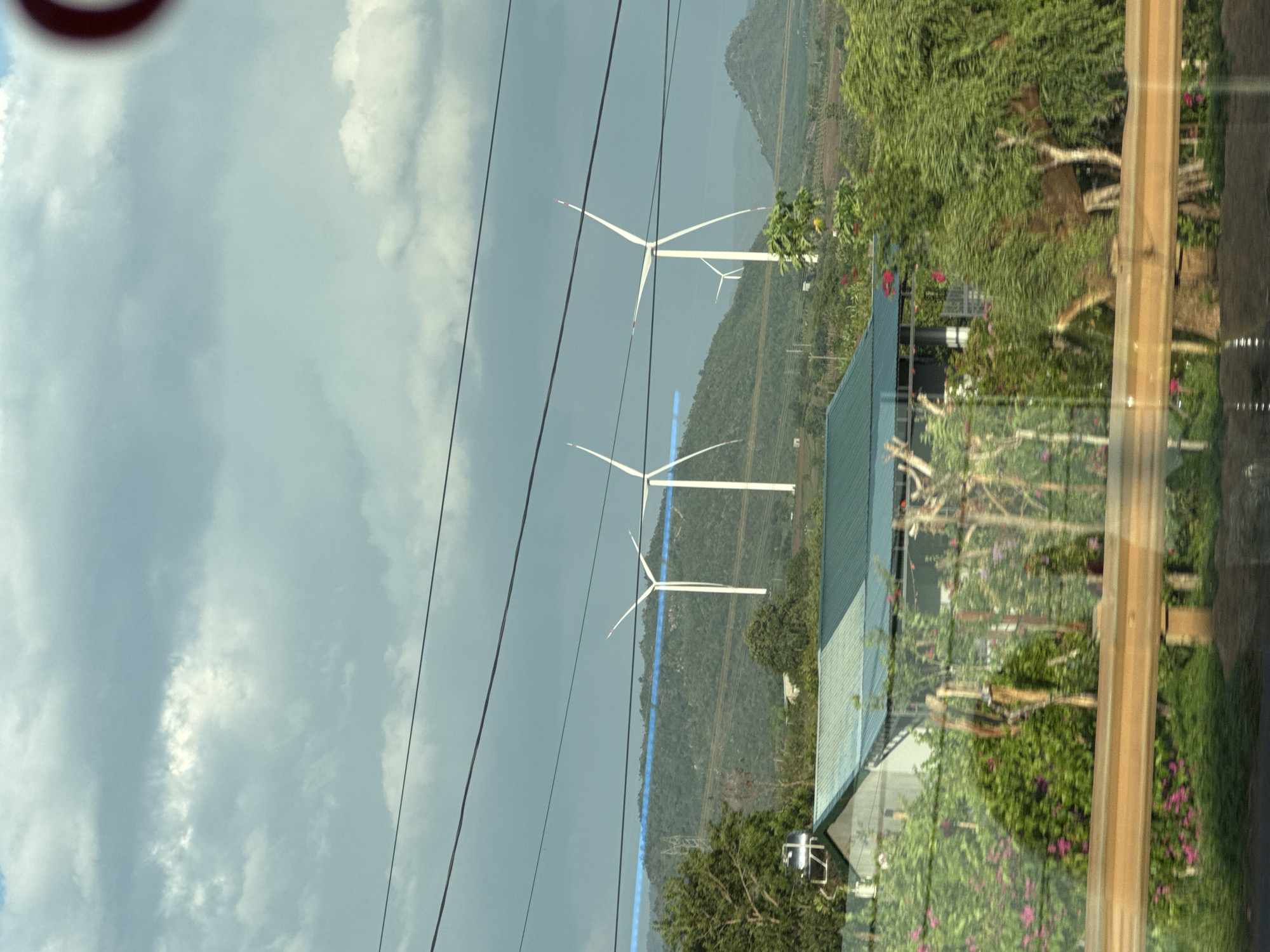 The image portrays a daytime landscape in Thôn 4, Vietnam, under an overcast sky. In the middle ground, three prominent white wind turbines with distinctive red-tipped blades are situated on a series of verdant, undulating mountains, indicating a wind farm. A striking blue line, possibly a road or a cleared path, runs horizontally across the distant mountain range.
Multiple power lines stretch across the upper part of the frame. To the lower right, a portion of a building with a dark green, angled roof and white walls is visible, nestled among lush green trees and bushes adorned with pink flowers. A water tank is also present near the building. The extreme foreground on the right displays a light brown wooden frame, likely part of a window or a vehicle's interior, showing reflections of the outside scene.
No specific human activity is depicted. The weather is cloudy but bright, suggesting mid-day. The scene captures a harmonious blend of natural beauty and modern renewable energy infrastructure in a rural, mountainous setting. No text is visible in the image.