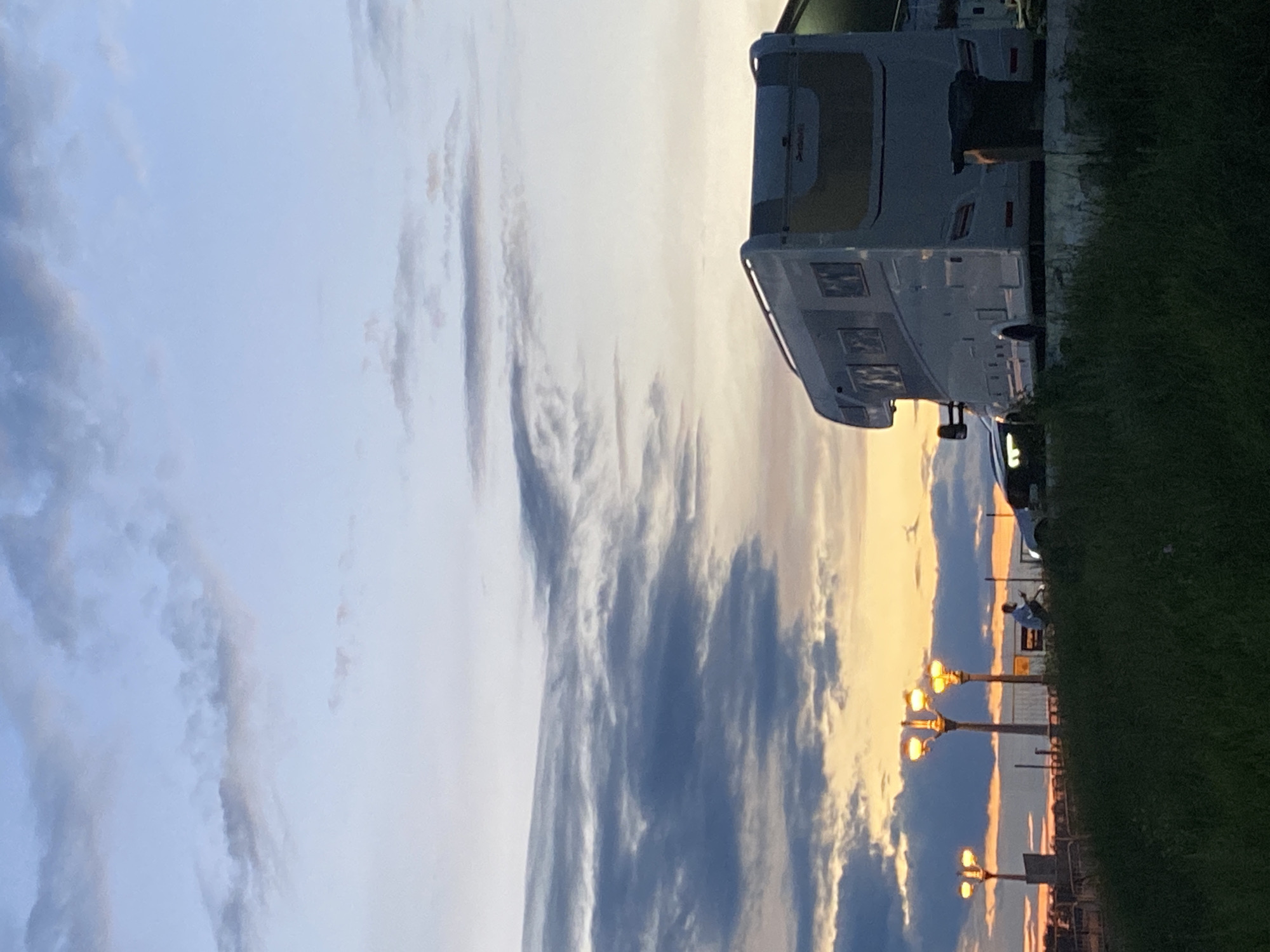The image depicts a scenic view at dusk in Novokuybyshevsk, Russia, with a motorhome parked on a slight incline. The sky is a gradient of pale blue and soft orange, with scattered clouds. In the foreground, tall green grass covers the ground. To the left of the motorhome, three lampposts with illuminated globes stand in a row. A person is seated on the ground near the lampposts, facing away from the camera and towards the horizon. A car is parked to the right of the person. The motorhome is large and white, with visible windows and a section of its rear visible.