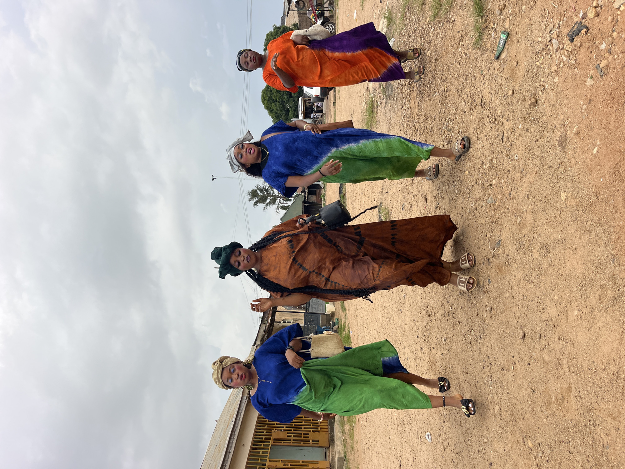 Four women are walking forward on a dirt path in an outdoor setting. The sky is overcast. The woman on the far left is wearing a blue dress with a green skirt, and a beige headwrap. She has blue eyeshadow and is wearing gold hoop earrings and black sandals. She carries a straw bag. The second woman from the left is wearing a long brown, tie-dye dress and a dark green headwrap. She has long braids and is wearing black sandals. She carries a black purse. The third woman from the left is wearing a blue and green tie-dye dress and a silver and white headwrap. She has bright blue eyeshadow and is wearing brown sandals. She carries a brown crossbody bag. The woman on the far right is wearing an orange and purple tie-dye dress and a black and white striped headwrap. She is wearing brown sandals. She carries a cream-colored bundle in her left arm. In the background, there are buildings, trees, a white car, and other vehicles. There is a green bottle and a piece of black plastic on the ground to the right of the path.