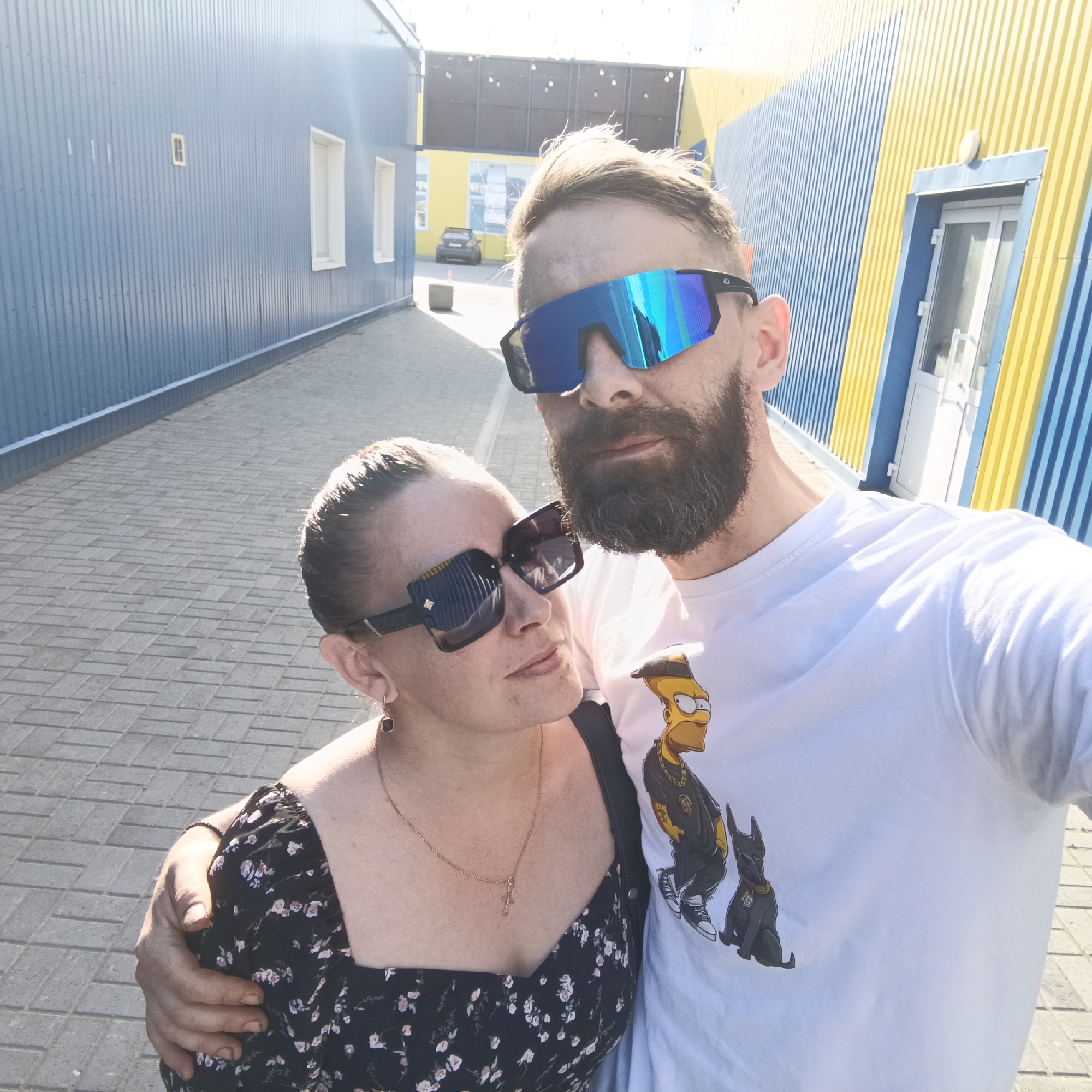 A selfie shows a man and a woman standing close together, with the man's arm around the woman. They are both wearing sunglasses. The man has a beard and is wearing a white t-shirt with a graphic of Bart Simpson in a black outfit and a dog. The woman is wearing a black floral dress and a necklace. The background shows a street with a blue and yellow building. Based on the metadata, the location is the city of Chaikovsky, Russia.