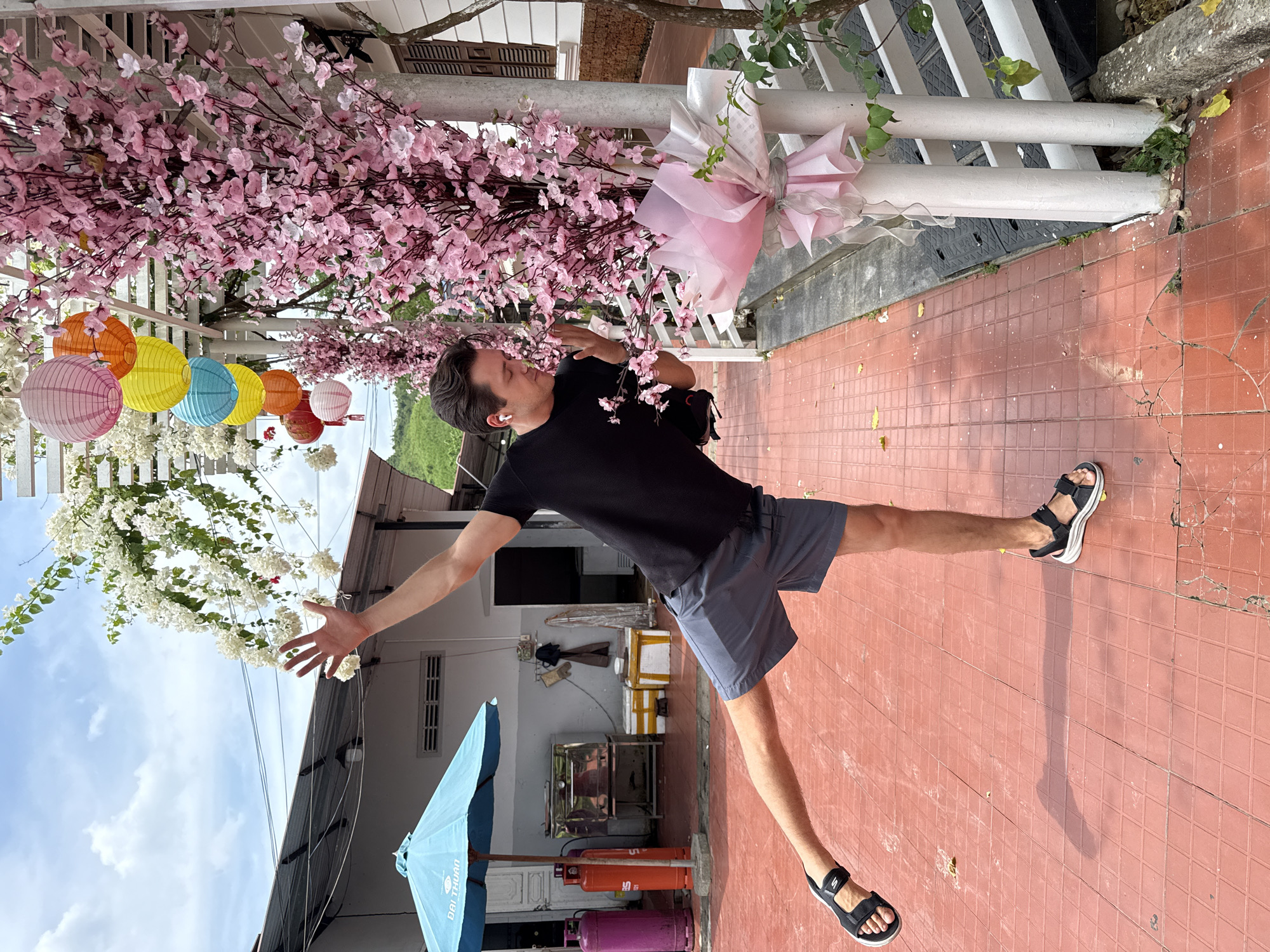 A man wearing a black shirt, gray shorts, and black sandals is in Nam Khánh Vĩnh, Vietnam. He is extending his arms in opposite directions and balancing on one leg on a red-tiled surface. The scene is decorated with pink and white floral arrangements, along with colorful lanterns. In the background, there is a blue umbrella, a building with an open front, and multiple propane tanks.