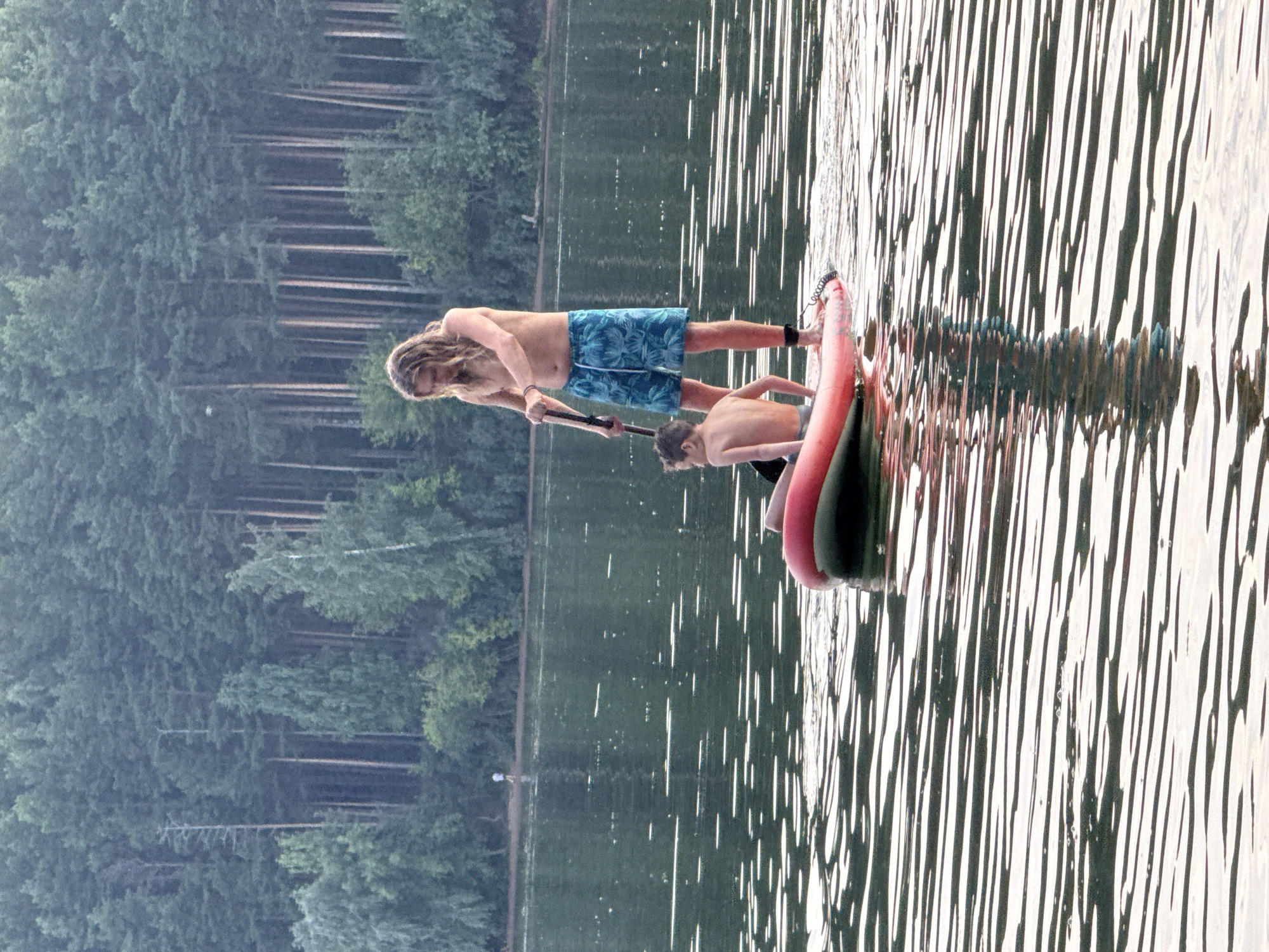 In a still body of water, two shirtless males are paddleboarding. The male in the foreground is younger and sits on the front of a red and grey paddleboard. The male behind him stands on the paddleboard and holds a paddle. He has long, light brown hair and is wearing blue patterned shorts. The paddleboard is positioned in the center of the frame, with the water creating a reflective surface. In the background, a dense forest of tall, green trees stretches across the horizon. The lighting suggests it is daytime, possibly late afternoon or early morning due to the soft light.