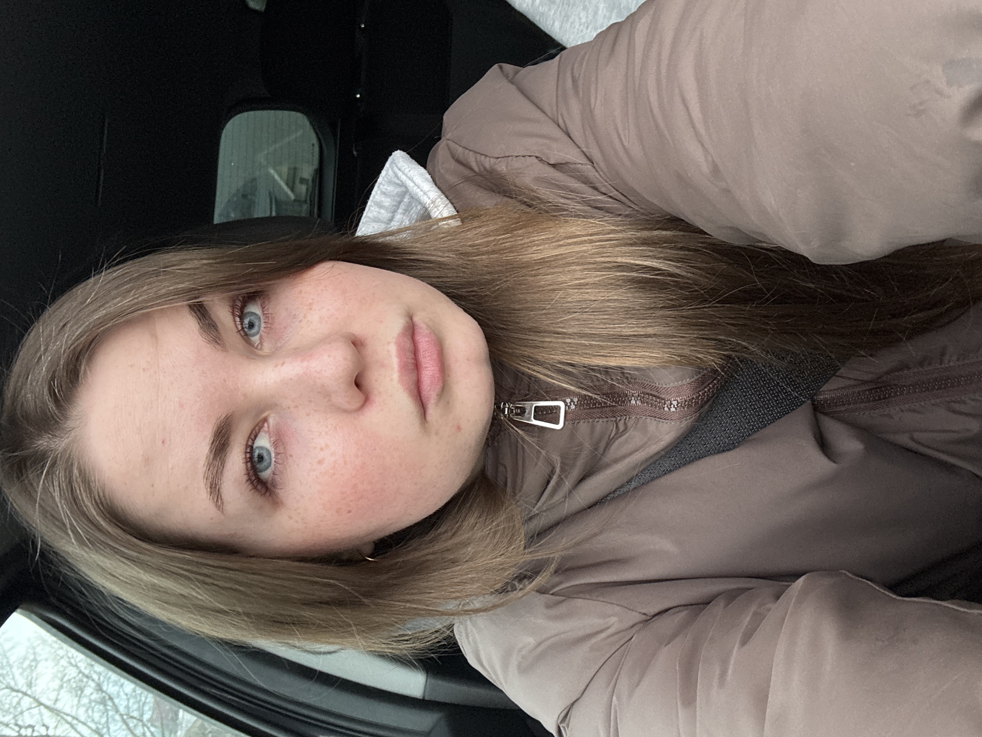 A woman is taking a selfie inside a car. She has light brown hair, blue eyes, and a scattering of freckles on her face. She is wearing a brown jacket and a grey hooded sweatshirt underneath. A seatbelt is visible across her torso. The interior of the car is dark, and the background visible through the car window is blurred, possibly indicating a street scene in Chaikovsky, Russia.