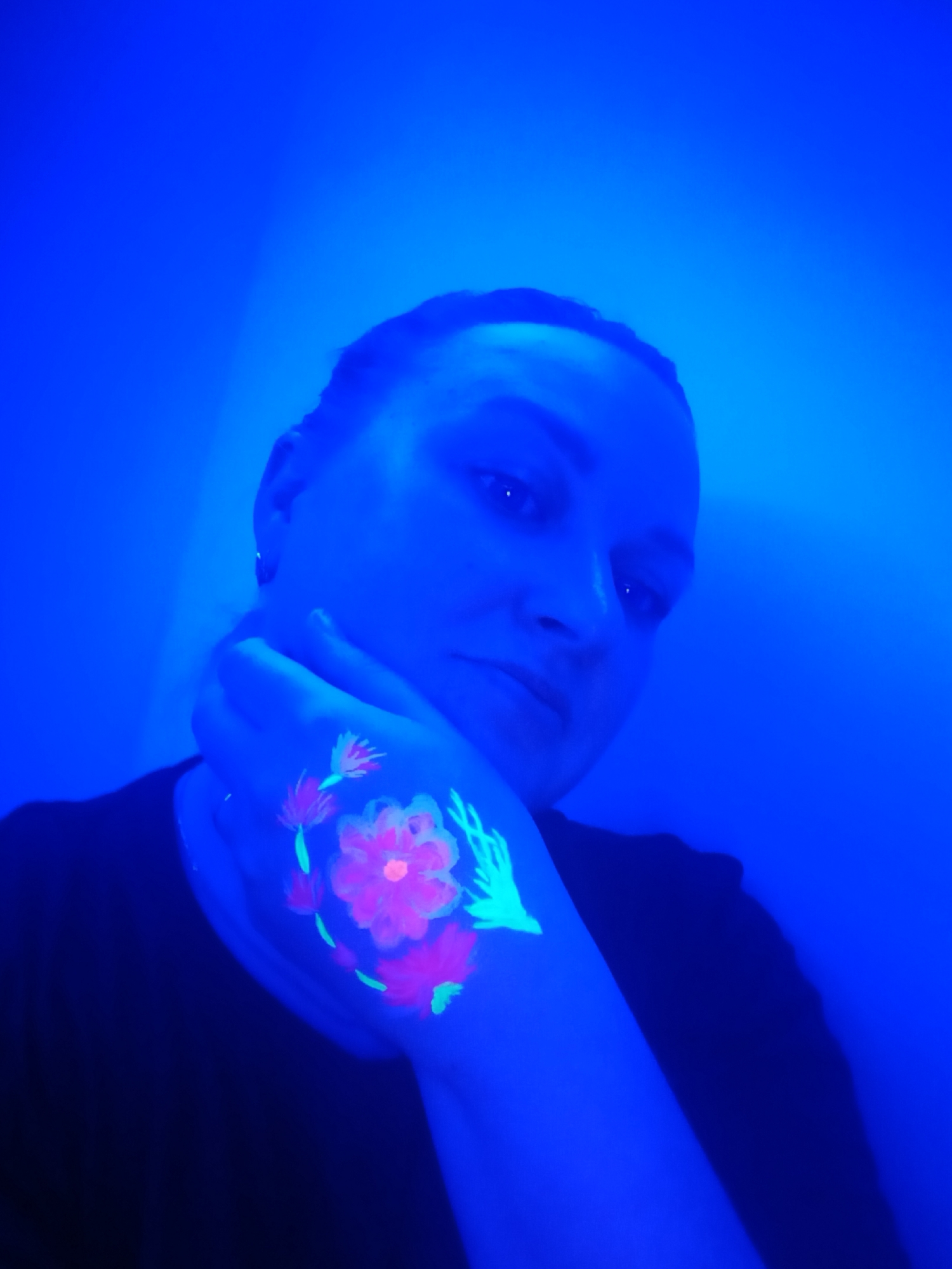 This media file features a person, likely an adult female with short hair, captured in an indoor setting in Chaikovsky, Russia, under strong blue ultraviolet (UV) lighting. The subject is wearing a dark top and holds her left hand near her face, looking towards the camera with a neutral or slightly contemplative expression.
The most notable detail is the intricate body art painted on her left hand and forearm, which fluoresces vibrantly under the UV light. The designs primarily consist of floral patterns rendered in bright neon pink and reddish hues for the blossoms, complemented by glowing green elements that resemble leaves or abstract stalks. The blue light permeates the entire scene, casting a deep blue tone over the subject's skin and creating a uniform, solid blue background, likely a wall. This specialized lighting condition suggests a night-time event, a club, an art installation, or a performance setting. No visible text is present in the image. The unique lighting creates an artistic and somewhat mysterious atmosphere.