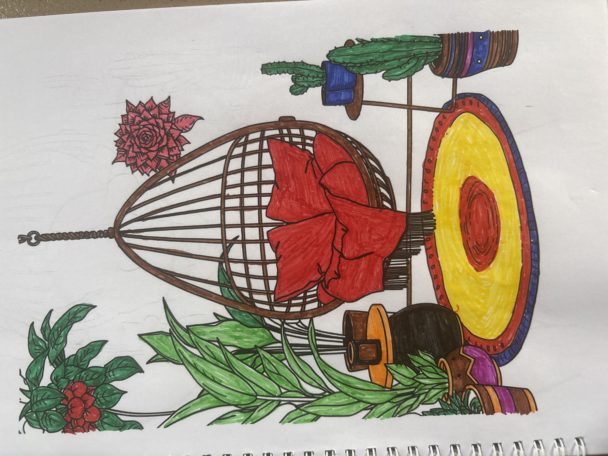 This is a colored drawing on white paper, depicting a bohemian-style indoor scene with plants and a hanging chair. On the left, there are two potted plants with large green leaves and a cluster of red flowers. A small table with an orange top and dark base sits in front of them, holding a small brown pot. In the center, a wicker hanging chair is suspended from the ceiling by a rope. The chair contains two red cushions. A colorful oval rug with red, yellow, and blue sections is placed on the floor beneath the chair. To the right of the chair, a small round table holds two blue pots with cacti. Another potted cactus with spiky green leaves is placed on the floor to the right of the table. The drawing is done in a somewhat childlike style with bold, flat colors. There are no people or animals present. The background is white, suggesting an indoor setting. The image does not contain any elements that would indicate a specific location in the city of Lafia, Nigeria.