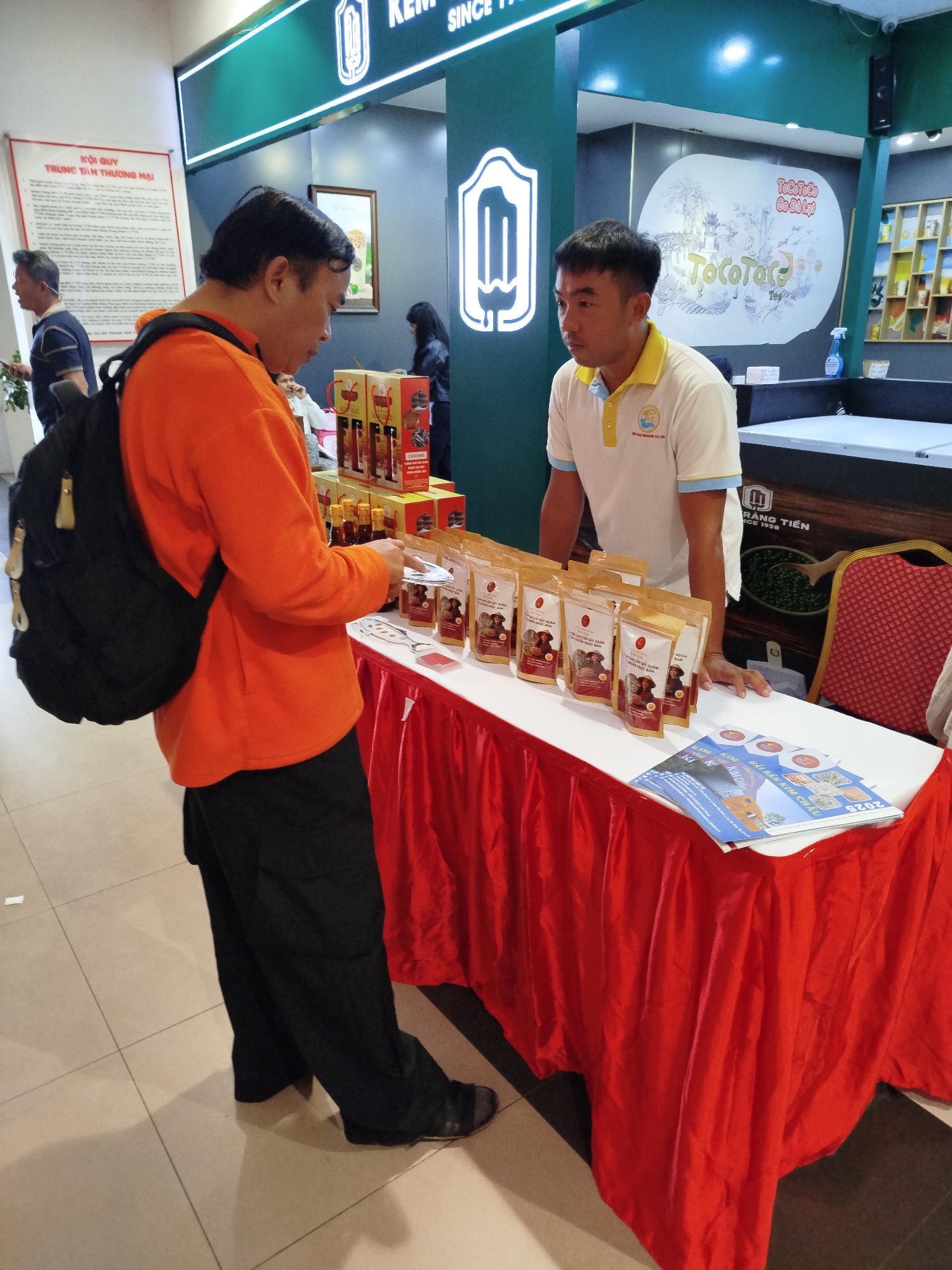 Two men are interacting at a display table covered with a red cloth, likely at a commercial event or store in Da Lat, Vietnam. The man on the left, wearing an orange long-sleeved shirt and a black backpack, is looking down at items on the table. The man on the right, wearing a white polo shirt with a yellow collar, appears to be a vendor, standing behind the table and looking at the other man. The table displays several packets of what appear to be food products and some brochures. In the background, there are store shelves with various products and signage, including one that reads "TOCOTOCO." The overall environment suggests a retail or promotional setting.