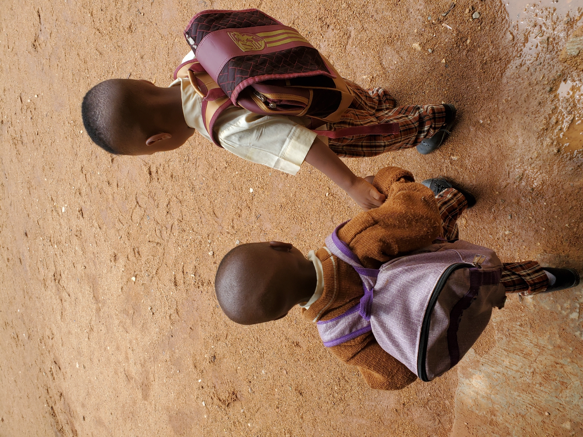 Two young children are captured from an overhead perspective, walking or standing on a reddish-brown dirt path under bright daylight. The ground is unpaved and sandy, with a distinct muddy puddle visible on the right, suggesting recent rain or collected water.
Both children appear to be wearing school uniforms and carrying backpacks. The child in the foreground is dressed in a light-colored (cream/beige) shirt, plaid trousers in shades of brown, orange, and red, and dark shoes or boots. They wear a maroon backpack featuring a quilted pattern, light brown straps, and a decorative gold-colored emblem or crest on the main flap.
The child positioned slightly behind and to the left wears a brown knitted sweater with prominent purple striped accents, plaid trousers (similar to the first child's), and dark shoes or boots. This child carries a light lavender or purple backpack with a dark zipper.
Both children have short, dark hair. Their posture suggests forward movement, likely on a journey to or from school. The reddish-brown soil and unpaved setting are characteristic of outdoor environments in areas like Jos, Nigeria. No discernible text is visible on the backpacks, only a decorative emblem.