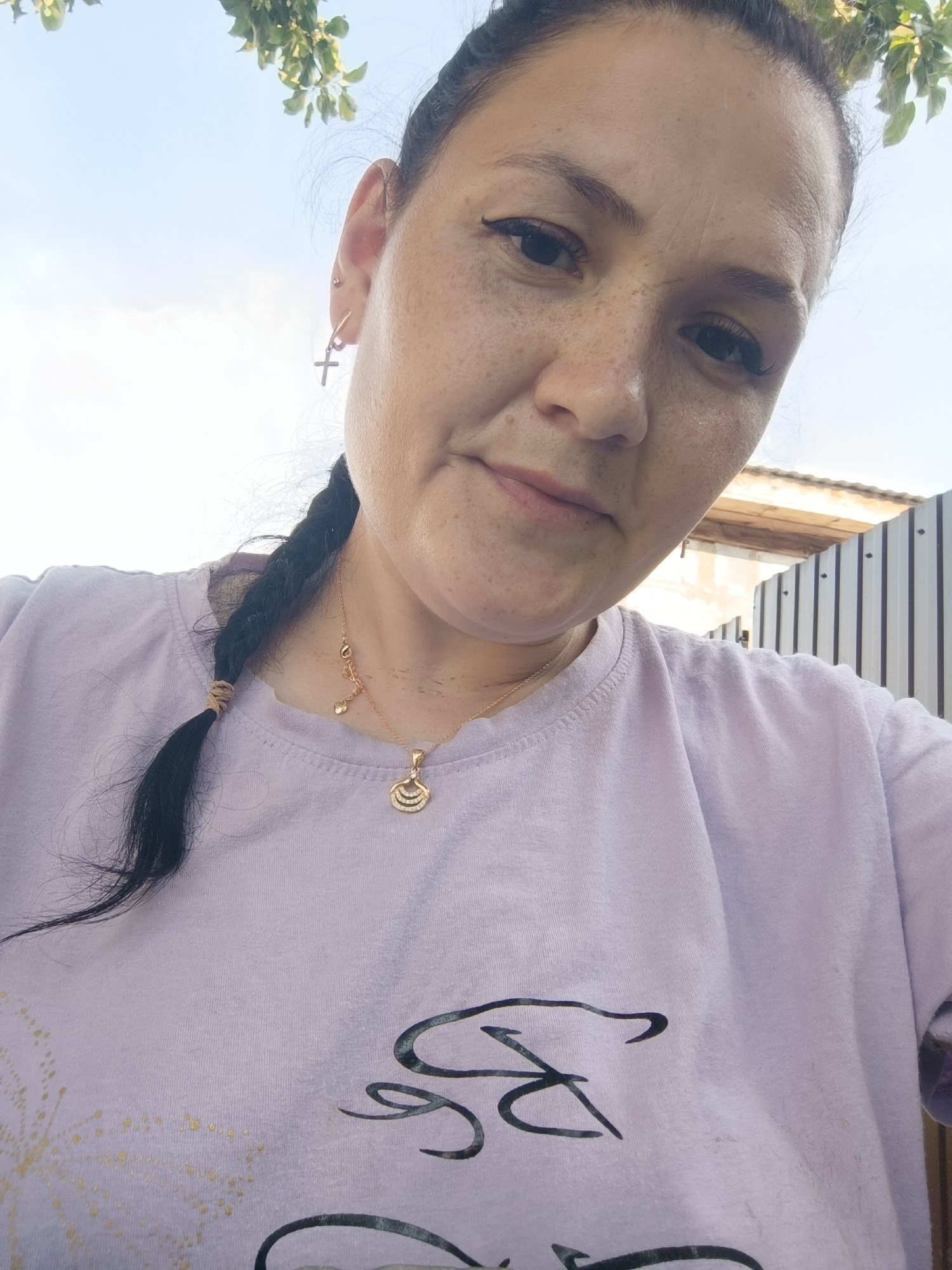 A woman with dark hair braided to her left, wearing a light purple t-shirt, is pictured from the chest up. She has a cross earring in her left ear and a gold necklace with two pendants, one of which is a shell-like design with three horizontal lines, rests on her shirt. She has freckles across her nose and cheeks and is looking slightly down and to her right. Behind her, a light blue sky is visible, with some green foliage from a tree in the upper left corner. To the right, a corrugated metal fence is present. The image is taken outdoors on a sunny day. There is no indication of the specific location in the city of Karsha, Russia.