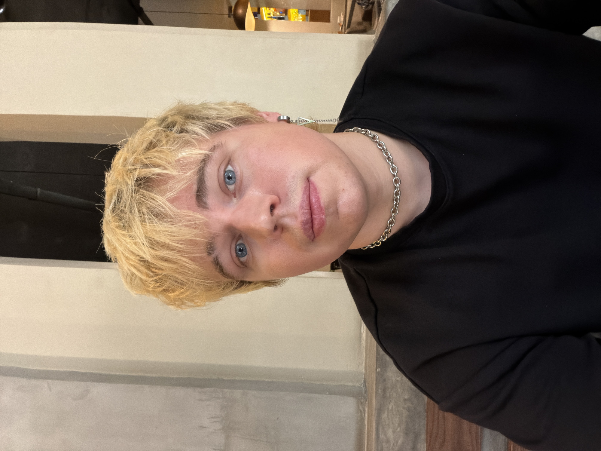 A headshot captures an individual with light blonde, shaggy hair and blue eyes, looking directly at the camera with a neutral expression. The person displays fair skin with some pink undertones. They are dressed in a plain black crew-neck top, accessorized with a silver-colored link chain necklace. A silver-toned ear cuff, featuring a triangular dangle and a chain, is visible on their right ear (viewer's left). The background reveals an indoor setting with light-colored walls, composed of a smooth beige section and a textured light gray concrete-like area. To the person's right, a darker interior space with an illuminated background element, possibly a bar or shelving, and a visible ceiling light fixture is present. A wooden surface is partially visible in the lower-left foreground. The scene is situated in Thủ Đức, Vietnam.