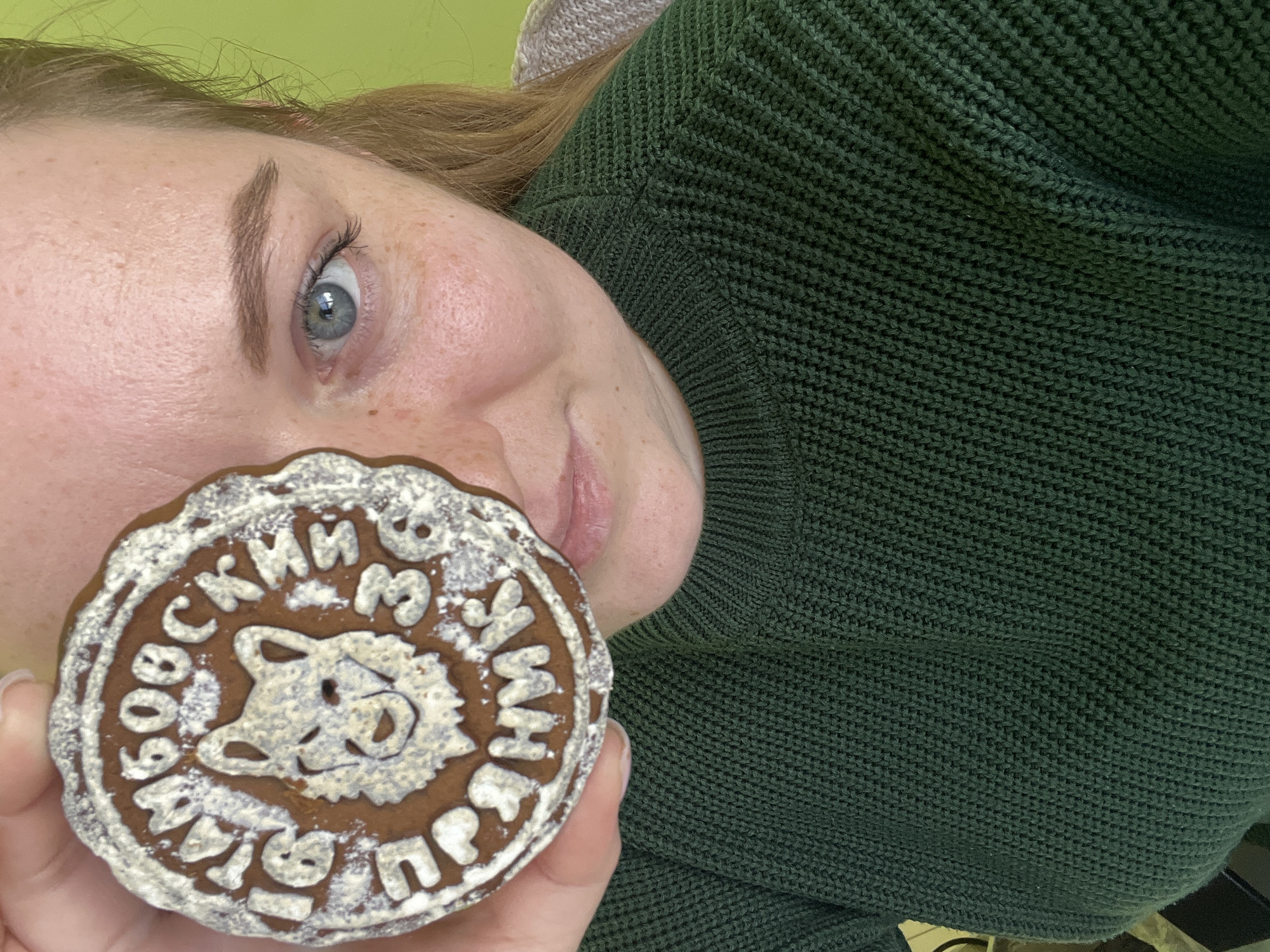 A person is holding a gingerbread cookie close to their face. The cookie is round, brown, and decorated with white icing. In the center of the cookie is a wolf's head, and around the edge are Russian words, including the name "Клин" (Klin). Given the Russian inscription on the cookie mentioning "Клин", it's reasonable to assume the location is Klin, Russia. The person has light skin, freckles, and blue eyes, and is wearing a dark green sweater. The background is a light green wall. The photo is taken at a close angle.
