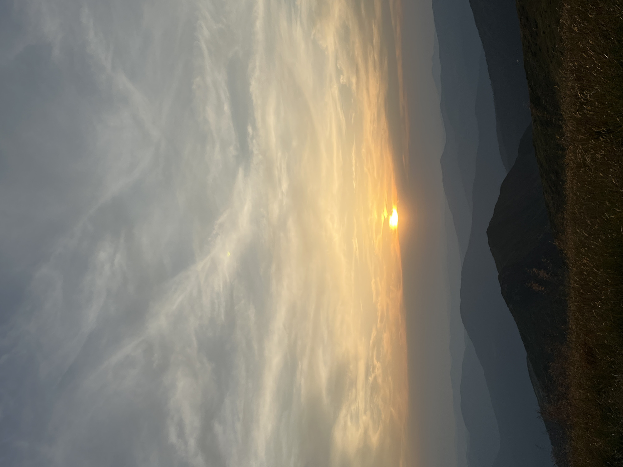 This is a vertical, eye-level, medium shot of a mountain range at sunset. The sun is a small, bright orb just above the horizon, casting a warm orange glow across the sky and illuminating wispy clouds. The foreground is dominated by dry, golden grass. In the mid-ground and background, several layers of mountains recede into the distance, their dark silhouettes creating a sense of depth. The image appears to be taken from a high vantage point, possibly a hilltop or mountain peak, overlooking the Carpathian Mountains in the area of Luhy, Ukraine. There are no people or animals visible in the frame. The environment is natural and mountainous, characterized by rolling hills, distant peaks, and a vast sky.