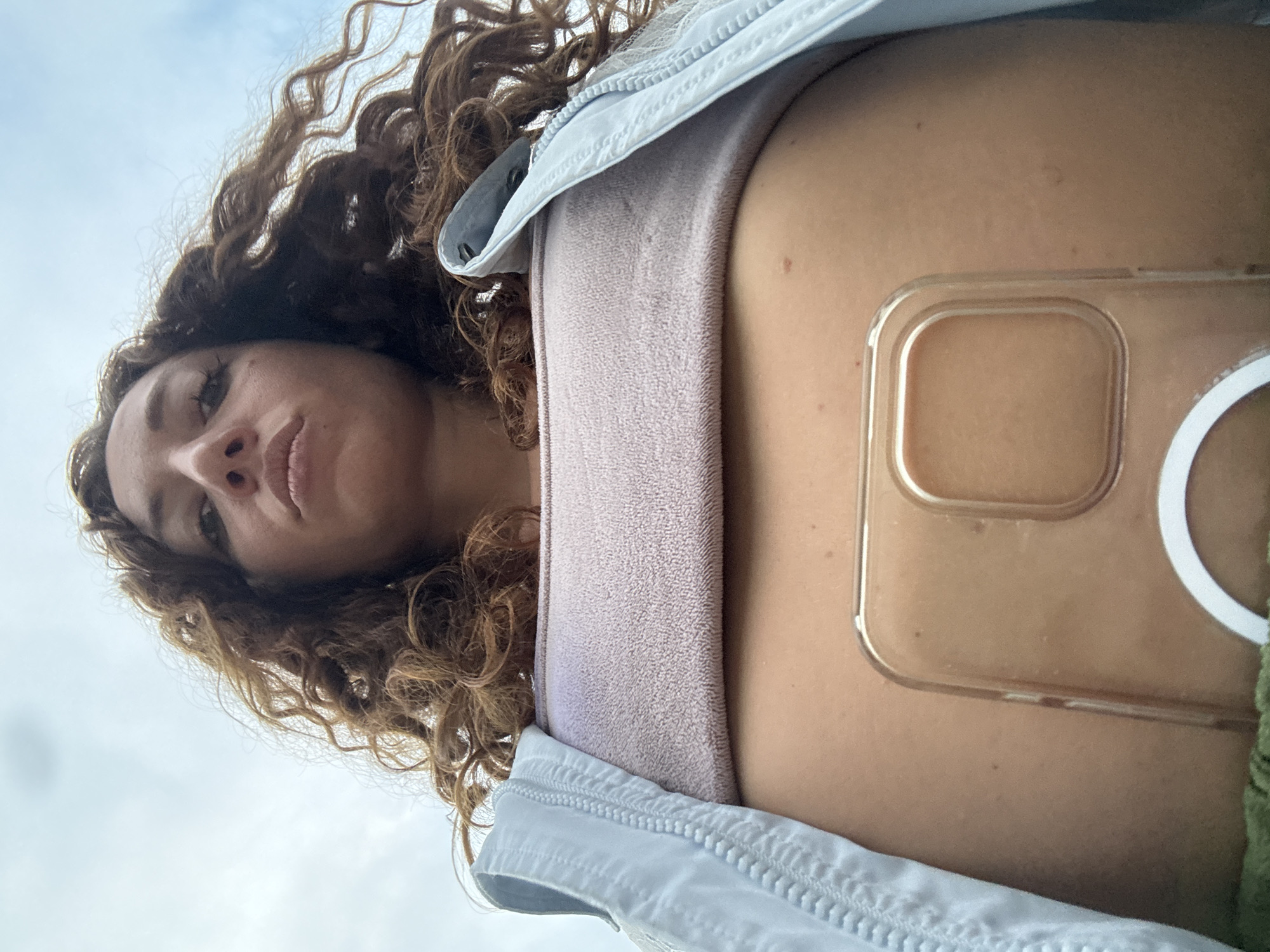 A low-angle, close-up shot captures a woman with curly, auburn hair and fair skin looking directly at the camera. She is wearing a light blue jacket over a light purple, textured bandeau top. A clear phone case with a white ring is placed on her stomach. The background consists of a cloudy sky. The image is taken outdoors, possibly in an urban or semi-urban environment, though no specific city details like Chiềng Châu, Vietnam, are evident.