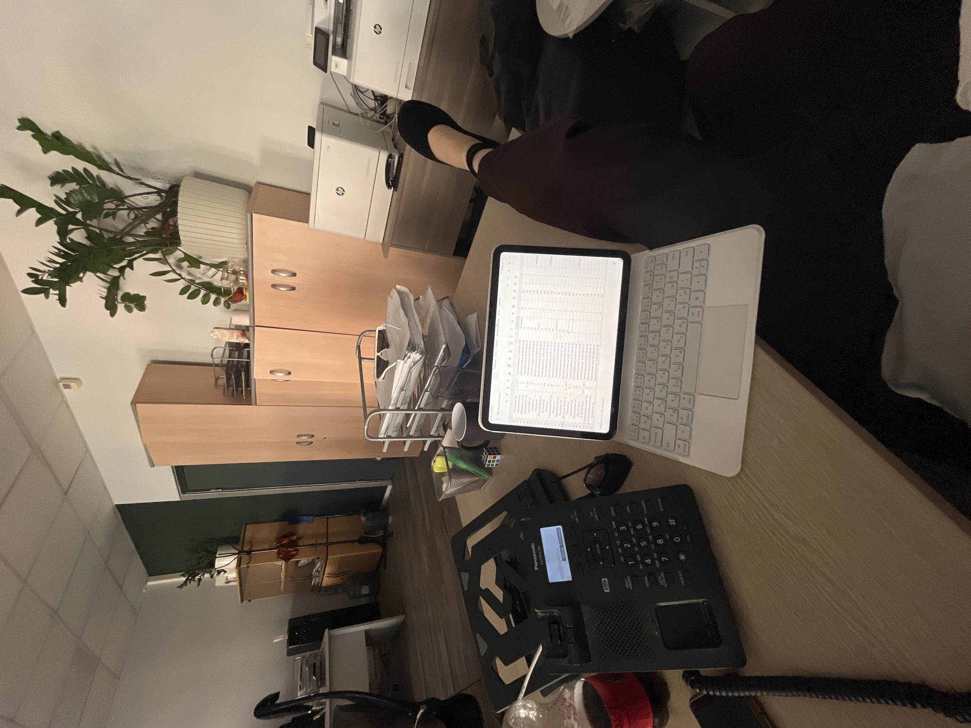 Here is a description of the image:
The image shows a desk in what appears to be an office setting. A person is seated at the desk, with their lower legs visible. The desk is cluttered with various items, including a tablet with a keyboard, a desk phone, a container with writing utensils, and stacks of paper in a wire organizer. There is also a plant on top of a cabinet beside the desk. The room appears to have standard office furniture and equipment. There is no evidence to confirm the location is in Moscow, Russia.