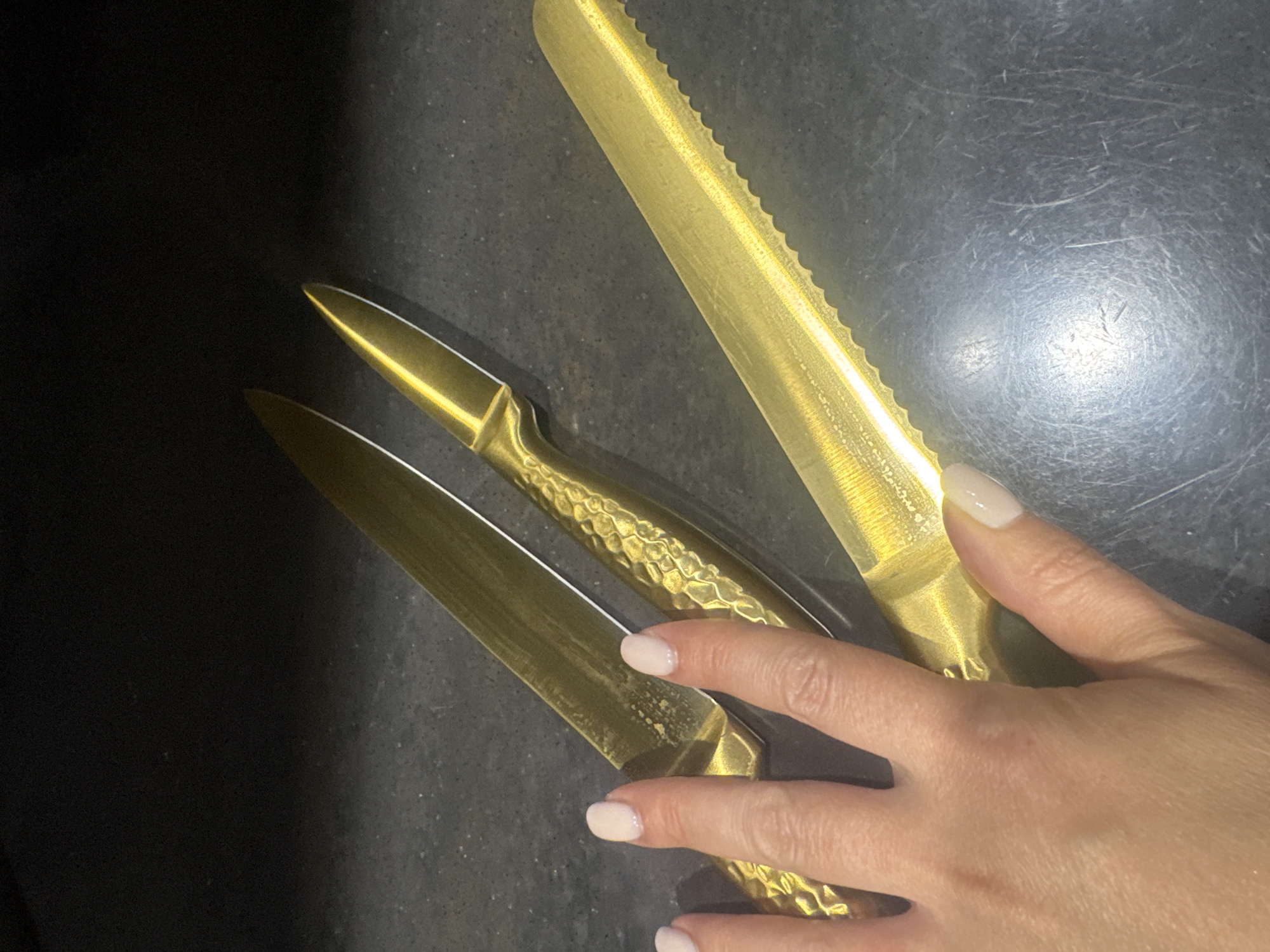 This media file shows a close-up overhead view of three gold-colored knives with metallic handles arranged on a dark, textured surface, possibly a countertop. Two of the knives have smooth, faceted blades, while the third, positioned diagonally across the frame, has a serrated edge, suggesting it's a bread knife or cake knife. A hand with light-colored, manicured nails is positioned on the right side of the frame, interacting with the handles of two of the knives. The lighting appears to be artificial, creating highlights and reflections on the metallic surfaces of the knives. The background is dark and out of focus, with some subtle texture visible in the surface the knives are resting on. There is no visible text in the image. The context suggests a domestic or professional kitchen setting.