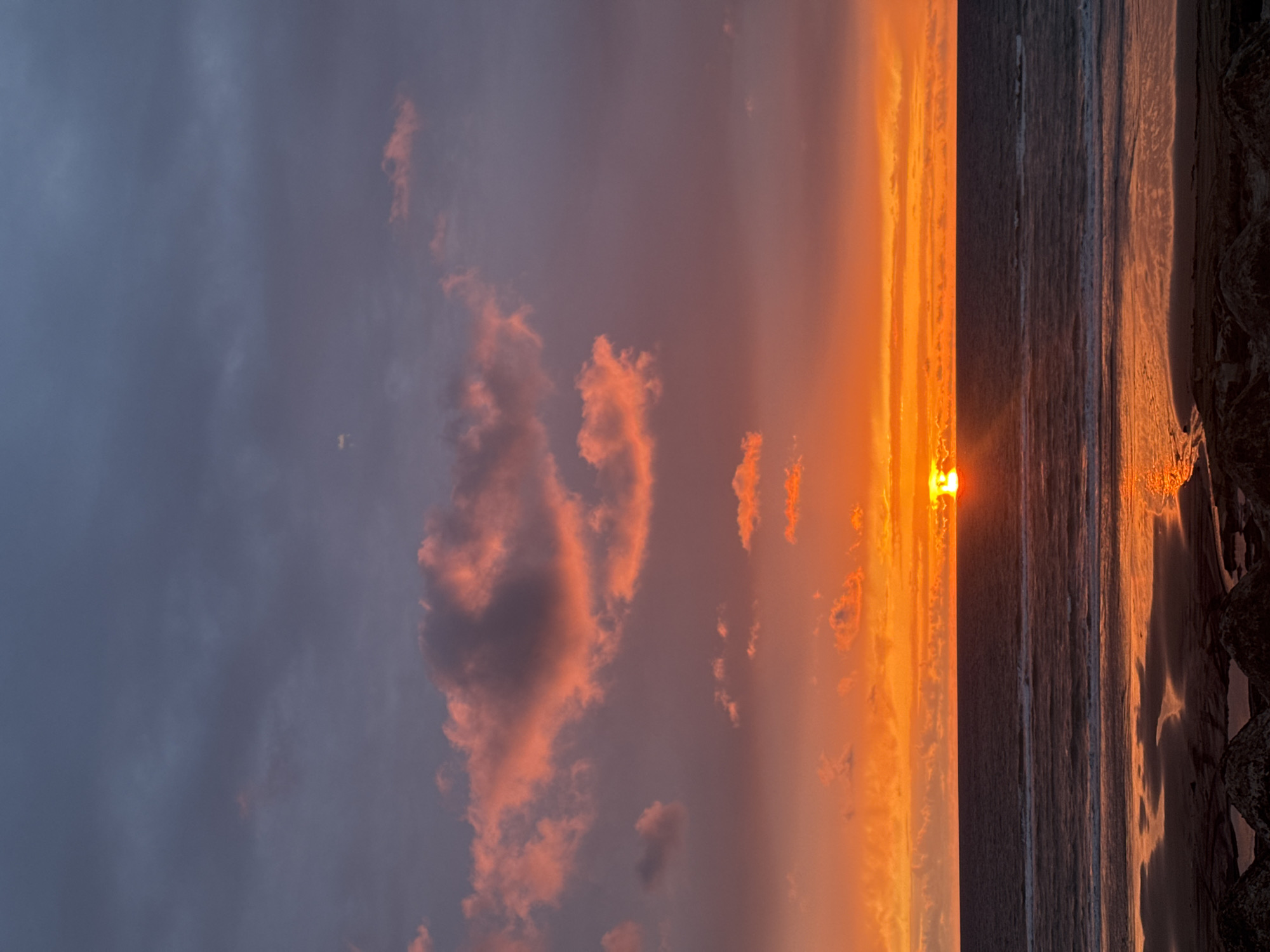 The image captures a vibrant sunset over the ocean, likely taken in Soulac-sur-Mer, France. The sky transitions from a dark, cloudy gray on the left to intense shades of orange and yellow towards the horizon. Clouds are scattered throughout the sky, some tinged with pink by the setting sun. The sun itself is positioned near the horizon, appearing as a bright, almost blinding orb. The ocean stretches out to meet the horizon, with subtle waves visible. The beach is dark and wet, reflecting the colors of the sky. Rocks are visible in the bottom right corner of the image. The orientation of the picture appears vertical, but the picture is sideways.