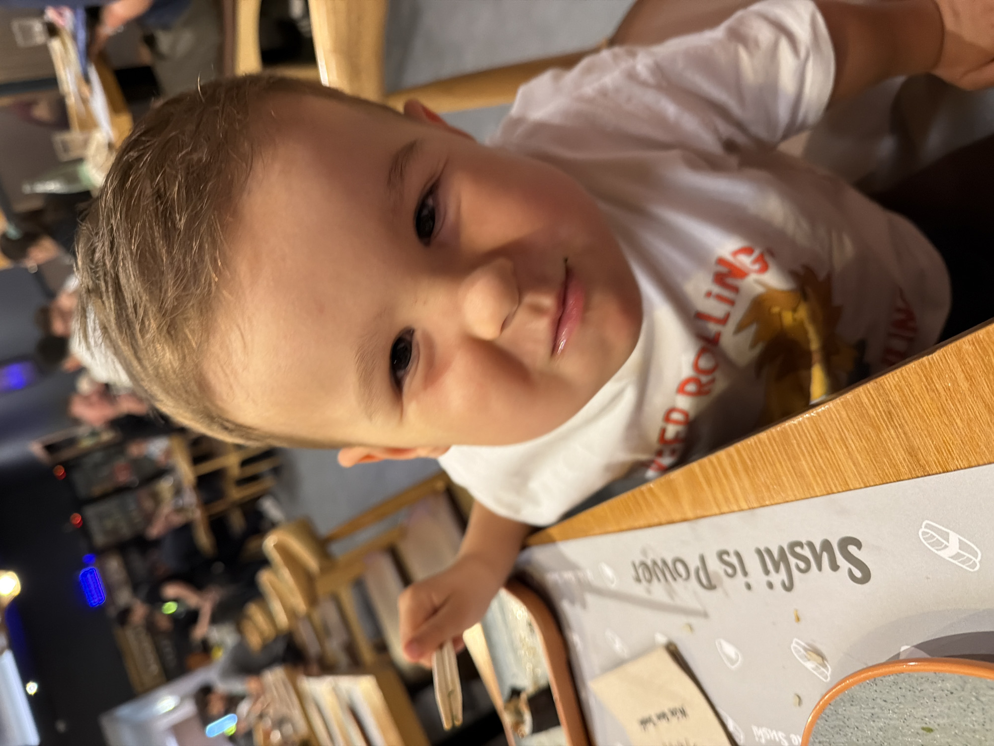 A close-up, eye-level shot shows a young boy with fair skin and light brown hair smiling directly at the camera. He is wearing a white t-shirt with red lettering that appears to say "RED ROLLING" and depicts a cartoonish yellow bird or eagle. The boy is seated at a table in what appears to be a restaurant, likely in Barcelona, Spain, given the visible text "Sushi is Power" on a grey placemat, a common phrase in sushi establishments. His right hand is raised, holding a wooden chopstick. In front of him is a sushi plate with a piece of sushi. The background is blurred, showing other diners and the restaurant's interior, including wooden chairs and tables, and some illuminated signs. The lighting is dim with some bright spotlights.
