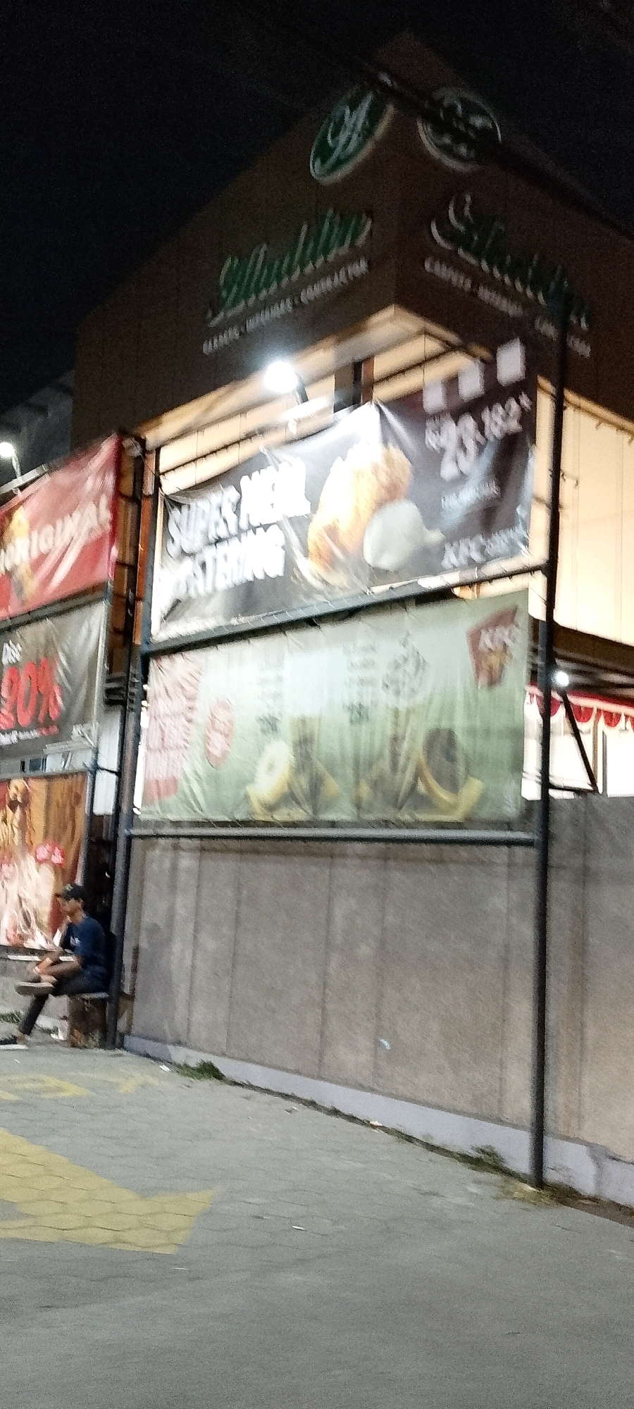 The image shows a street scene in Gunung Putri, Indonesia at night. In the background, there is a building with signs for "Madinah" and "KFC". A banner on the building advertises "Super Meal Catering". In the foreground, a person is sitting on a bench outside the building. The street is paved with interlocking concrete blocks and there is a yellow directional arrow painted on the ground. The lighting suggests an outdoor commercial area.