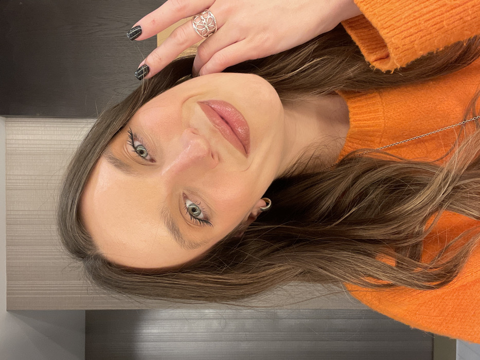 A woman in an orange sweater tilts her head, displaying a silver ring on her hand. She has long, wavy brown hair, fair skin, and subtle makeup highlighting her eyes and lips. Her nails are painted dark with a textured finish. A small silver earring is visible. The background includes a wood surface and a neutral-toned wall. The lighting is bright and even, showcasing the woman's features.