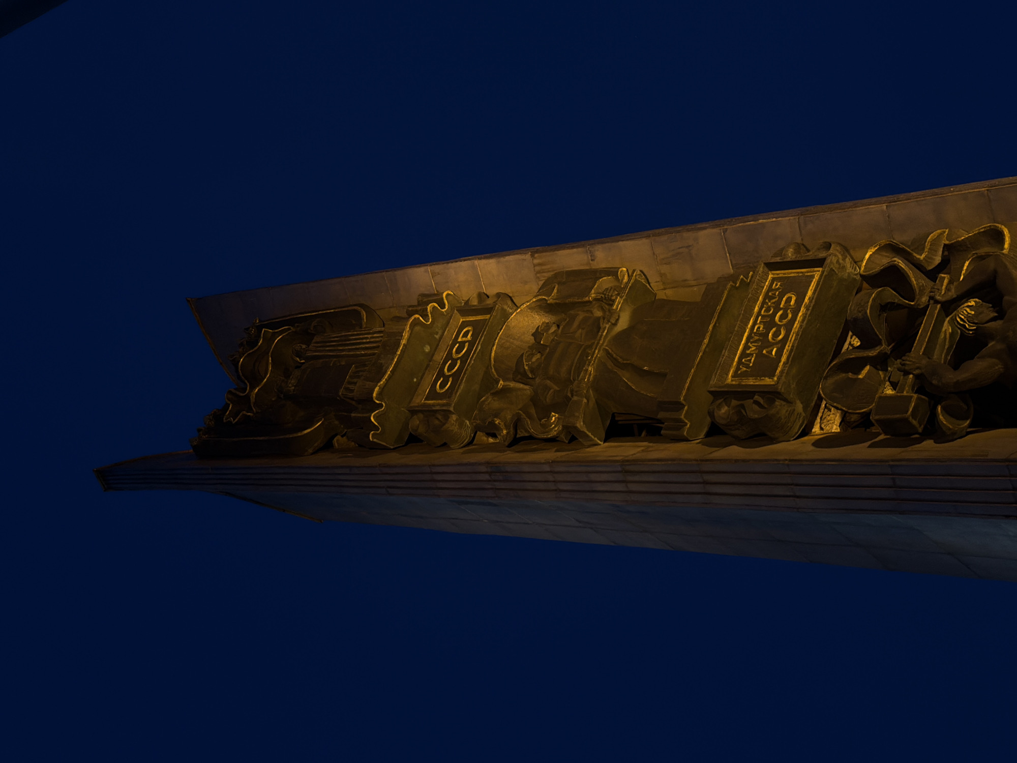 The image captures a segment of the "Friendship of Peoples" stele in Izhevsk, Russia, focusing on its monumental bronze relief at night. The relief, illuminated by artificial lighting, depicts stylized figures and symbols associated with the Udmurt Autonomous Soviet Socialist Republic. Inscriptions in Cyrillic script reading "CCCP" and "УДМУРТСКАЯ АССР" (Udmurt ASSR) are visible. The background is a dark, near-black night sky. The perspective is from a low angle.