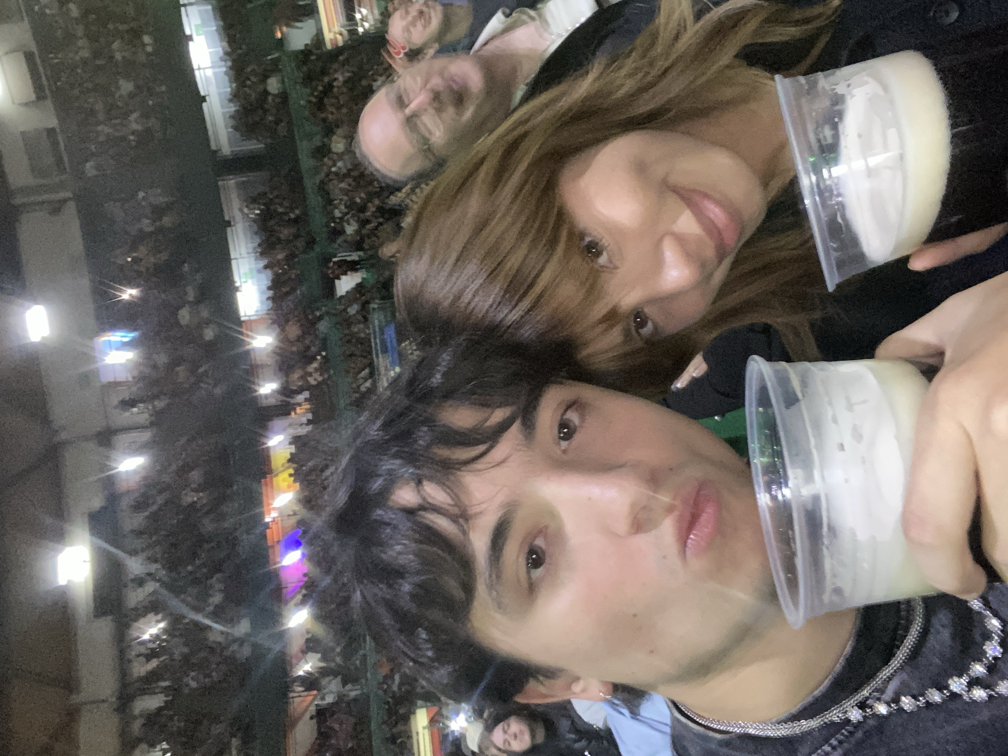 An indoor scene depicts two individuals in the foreground, appearing to take a selfie. A young man, with dark, curly hair and light skin, faces the camera with pursed lips, wearing a dark t-shirt and layered silver-toned necklaces. He holds a clear plastic cup containing a light-colored, frothy beverage. To his right, a young woman with shoulder-length brown hair and bangs smiles towards the camera, leaning slightly towards the man. She wears a dark jacket and holds a clear plastic cup with a darker, frothy beverage.
Behind the two subjects, a large crowd of spectators fills tiered seating sections of an indoor arena. An older man with glasses, wearing a light-colored shirt, is visible smiling in the middle-right background, positioned behind the young woman. Other indistinct faces are scattered throughout the background crowd. Bright stage lights in purple, blue, orange, and white hues are visible in the distant background, indicating a live event or concert. The overall lighting is dim, characteristic of an indoor entertainment venue.
The event is taking place at the Palacio de los Deportes, located in Mexico City, Mexico.