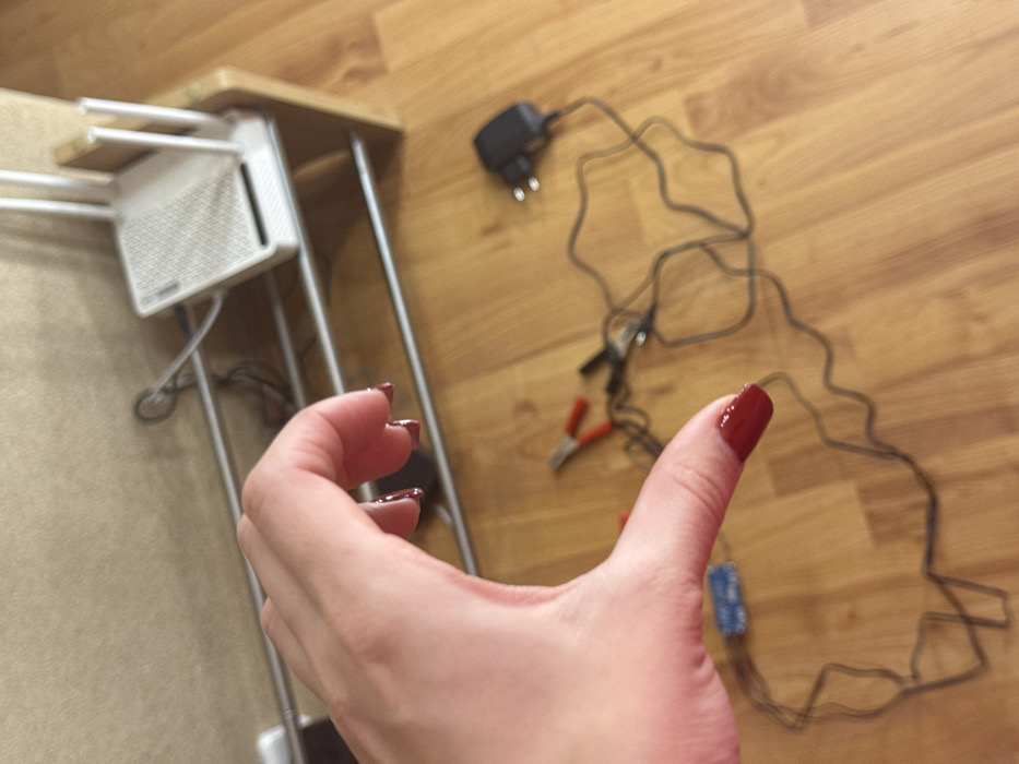 A hand gives a thumbs-up, showing a thumb with dark red nail polish. In the background, a wooden floor hosts a collection of electronic components including a black power adapter, wires, a circuit board, and alligator clips. Beside the hand, a wireless router with multiple antennas sits on a small, light-colored table or stand.