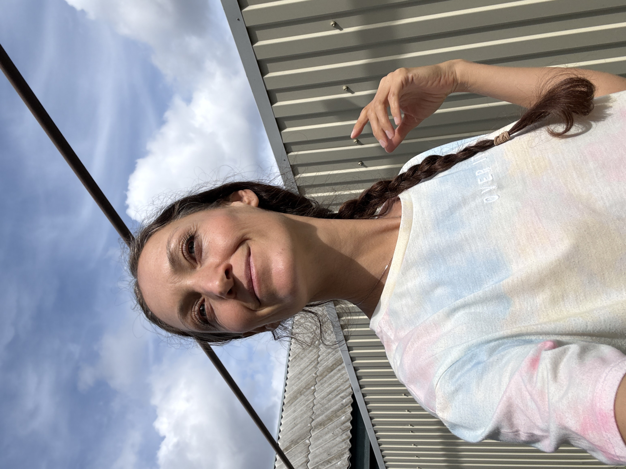 A woman with long brown hair braided in a single plait over her right shoulder smiles at the camera. She is wearing a t-shirt with a light tie-dye pattern in shades of pink, blue, and white. The background includes a cloudy sky and a corrugated metal structure. There is a pole in the upper left corner of the image. The location cannot be verified as Ulyanovsk, Russia, as there are no distinguishing features in the photo.