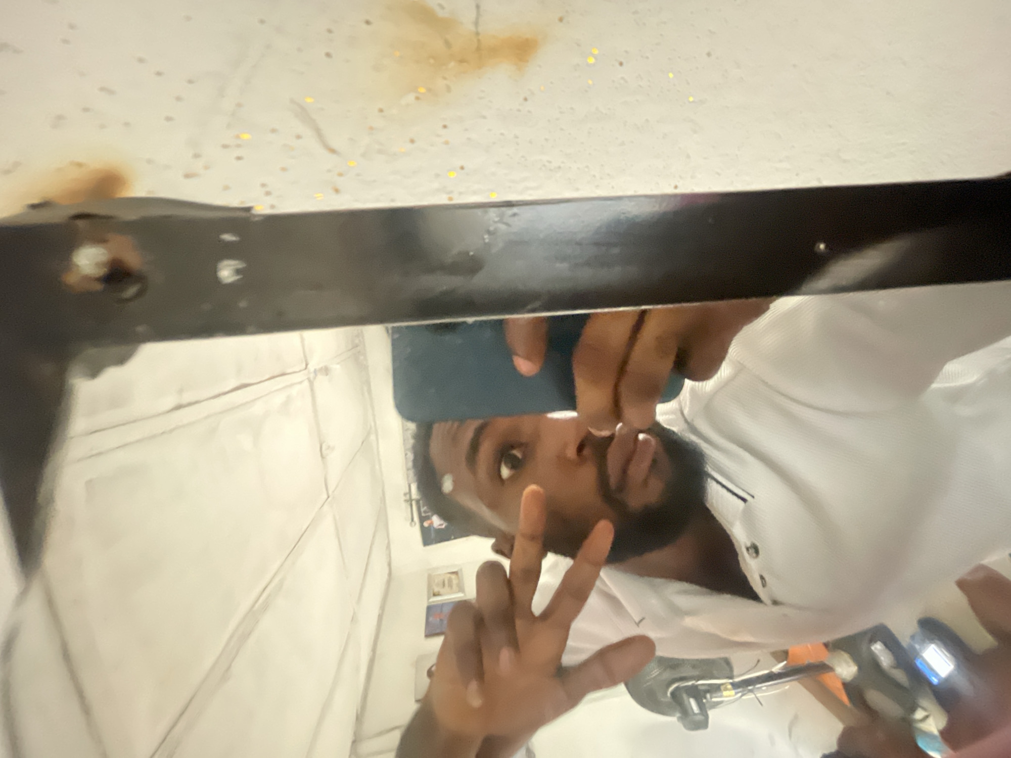 The image is a selfie taken in a mirror. A Black male with a beard is visible, holding a phone to take the picture. He is wearing a white collared shirt and making a peace sign. The reflection in the mirror reveals a tiled wall, likely of a bathroom. In the background, small framed pictures are visible. The top of the mirror frame is a dark color with some discoloration on the surface. The location is unknown and could not be described within Lokoja, Nigeria.
