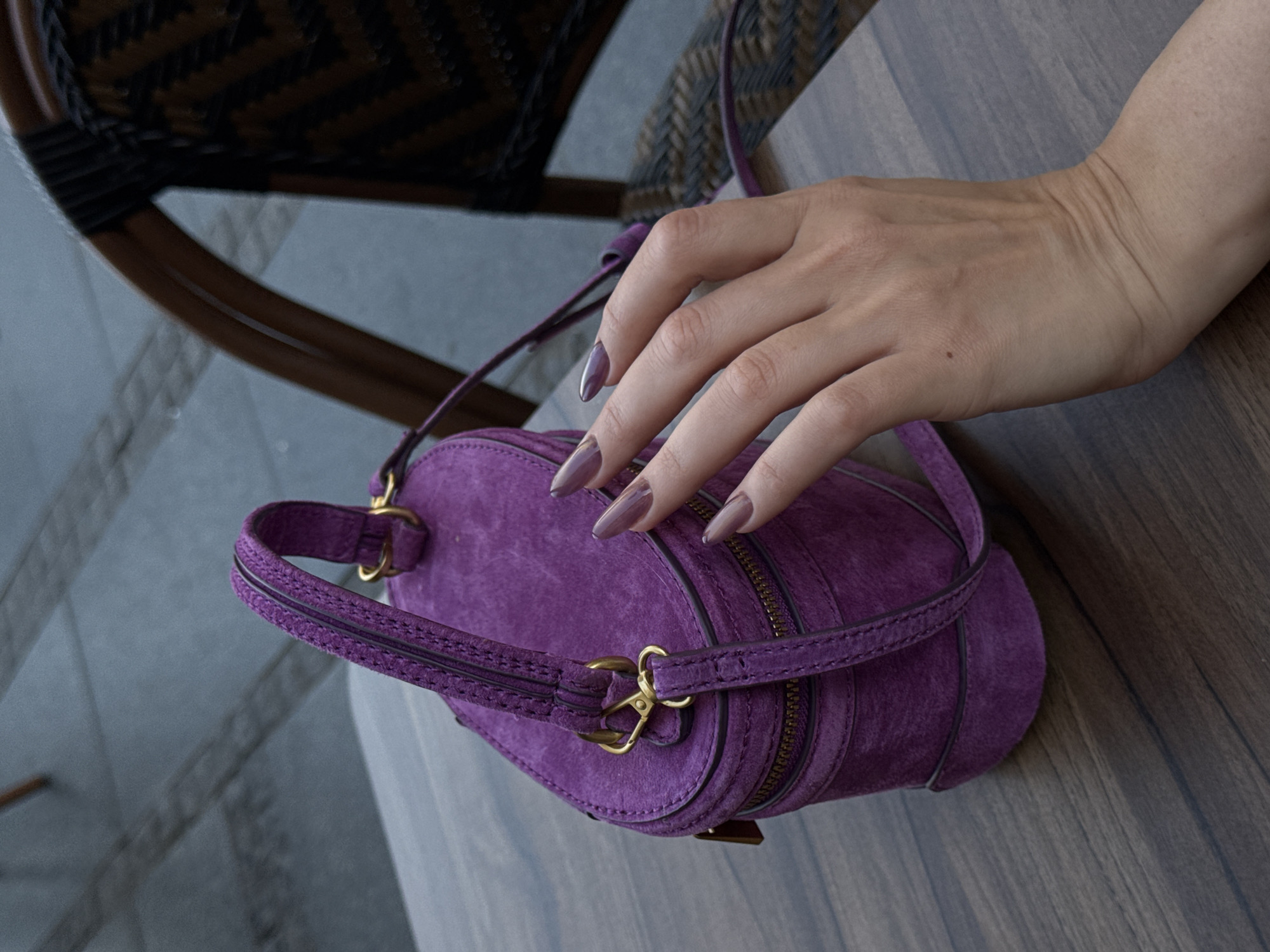 A hand is resting on a small, purple suede purse that is lying on a wooden surface. The purse has gold-colored hardware and a visible zipper. The hand has long, polished nails with a mauve color. A portion of a wicker chair is visible in the background. The location is Belgrade, Serbia.