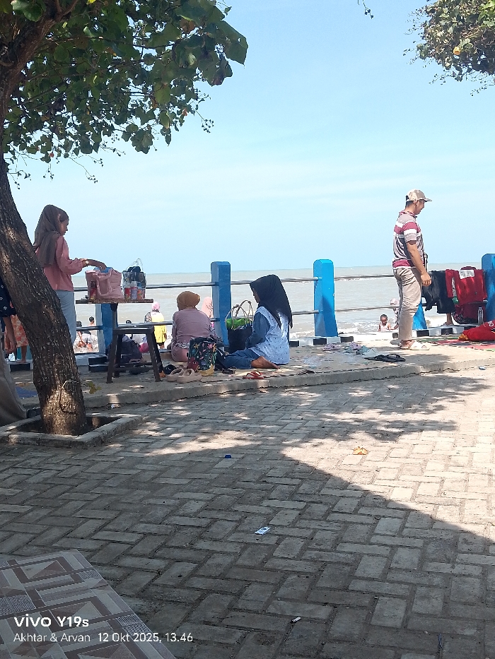 This image captures a bright, daytime coastal scene in Limbangan, Indonesia. The foreground features a light-colored interlocking paver walkway, partially shaded by the dense green foliage of a tree whose dark trunk anchors the left side of the frame. Beyond this walkway, a raised concrete platform with a blue and light blue painted metal railing overlooks the sea.
Six individuals are present. On the left, a female in a light brown hijab and pink long-sleeved shirt stands beside a two-tiered dark wooden table, which holds a light pink bag and other items. She is actively engaging with objects on the table. To her right, on the raised platform, a person wearing a light brown hijab and light-colored top is seated, partially turned away from the camera, facing the sea. Adjacent to them, a female in a black hijab and a light blue patterned long-sleeved top over blue jeans is also seated on the platform, oriented towards the sea, with a green bag and a floral backpack nearby. Multiple pairs of footwear, including platform sandals, red slippers, and black sandals, are scattered on the platform in this area. Further right, a male in a striped short-sleeved shirt, light pants, and a baseball cap stands with his back partially to the camera, looking towards the right and the sea. In the distant background, several indistinct figures are visible in the shallow ocean water. The sea exhibits light waves under a clear blue sky.