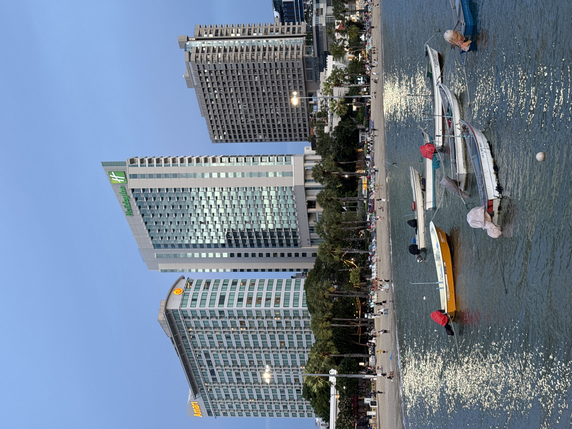 This image captures a picturesque view of Pattaya City, Thailand, specifically the beachfront area. In the foreground, several small boats are anchored in the calm, blue water, their reflections shimmering on the surface. The boats vary in color and design, with some appearing to be fishing boats or small leisure craft. One boat in the center is a striking orange color and is covered with a red tarp.
Beyond the boats, a sandy beach is visible, populated by numerous people. The beach is lined with palm trees and further back, several large buildings dominate the skyline. Among these are prominent hotels, including the Holiday Inn with its distinctive green logo and the Amari hotel. The architecture suggests a modern, developed urban setting. The sky above is a soft, twilight blue, indicating that the photograph was taken either at dusk or dawn. String lights are visible along the beachfront, suggesting a lively atmosphere and possibly the presence of restaurants or stalls. The overall scene conveys a sense of a bustling coastal city, a popular tourist destination.