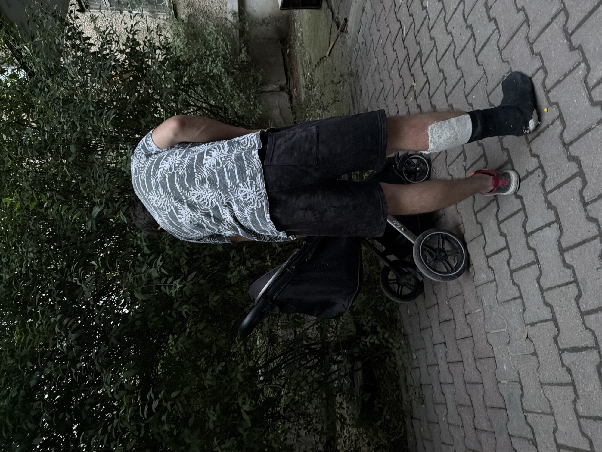 A person is standing on a paved walkway next to a stroller. The person is wearing black shorts and a grey t-shirt with a white palm leaf pattern. Their left leg is bandaged from the knee down and covered by a black boot. The right leg is visible and has a red and grey sneaker. The stroller is black and appears to be folded. In the background, there is lush green foliage and the corner of a building. The scene is outdoors, with the lighting suggesting it is late afternoon or early evening. The paving stones indicate an urban or suburban setting, possibly a park or residential area. The overall impression is of someone managing with an injury, possibly taking a child out for a walk. No visible text is present in the image. The location context provided is Shymkent, Kazakhstan.