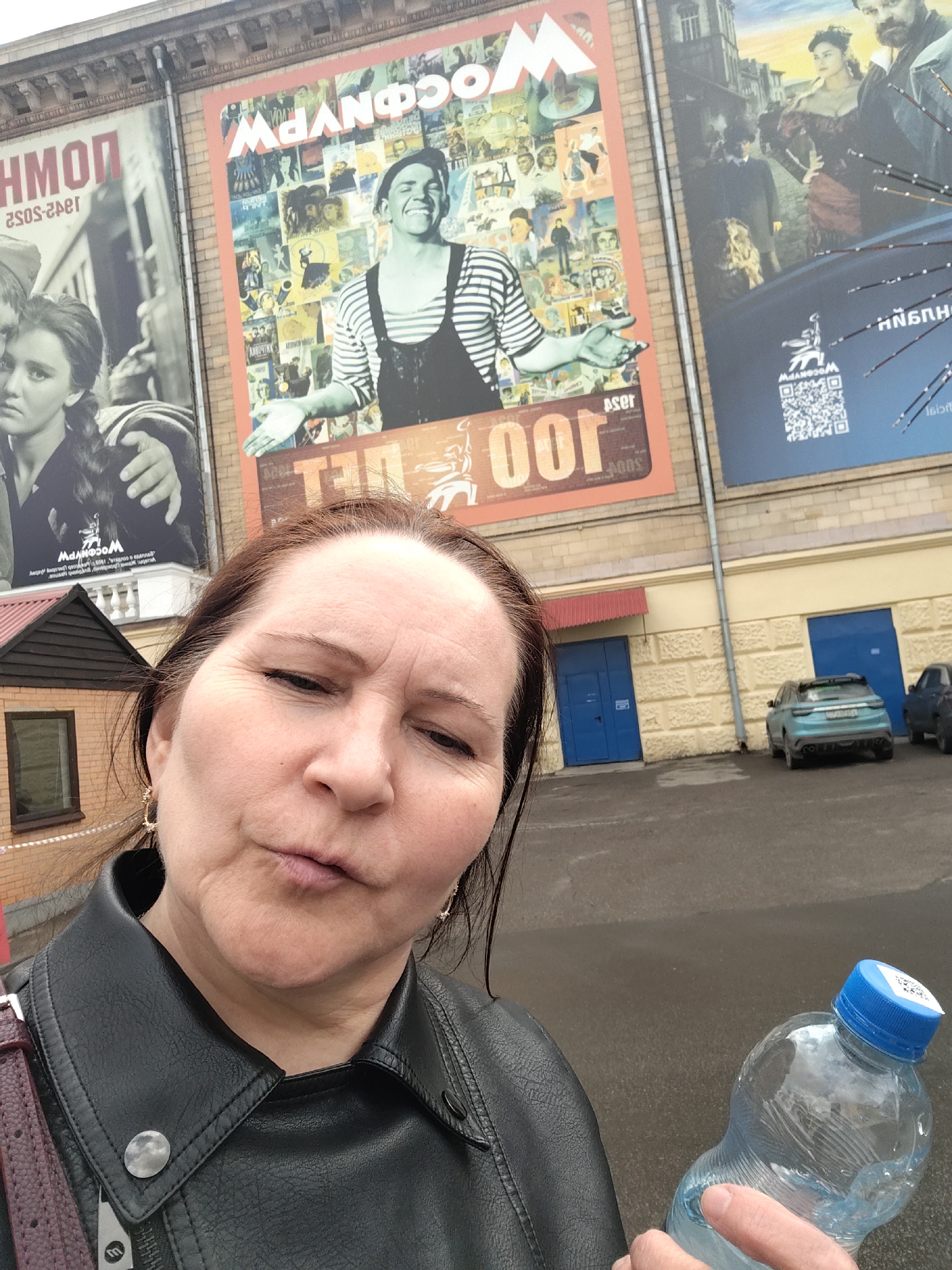 A woman takes a selfie in an outdoor setting, holding a clear plastic water bottle with a blue cap. She has dark hair, is wearing a black leather jacket with a silver-colored snap, and gold-colored earrings. Her expression is a slight pout or squint, looking towards the camera.
Behind her is a large building with a light-colored, textured facade. Dominating the background are three large murals or posters, typical of cinema advertising. The central and most prominent poster features a smiling man in a striped shirt and dark overalls, set against a collage of film stills and images. At the top of this poster, the word "МОСФИЛЬМ" (Mosfilm) is clearly visible, and the number "100" is prominently displayed below the man, suggesting a centennial celebration for the famous Russian film studio.
To the left, a partially visible poster shows a young woman with a braided hairstyle, rendered in black and white, and partially mirrored text that includes "КИНОП" (KINO P) and "1942-2052". To the right, another poster features figures in historical attire and also displays the "МОСФИЛЬМ" logo and a QR code.
In the mid-ground, a small brick structure with a brown roof and a window is visible to the left. Below the murals, the building has two blue doors. A paved area or parking lot extends from the building, where a light blue SUV-like car and another darker vehicle are parked. The ground appears slightly damp, and the lighting suggests an overcast daytime. The overall context, particularly the "Mosfilm" branding, places the scene in Moscow, Russia.