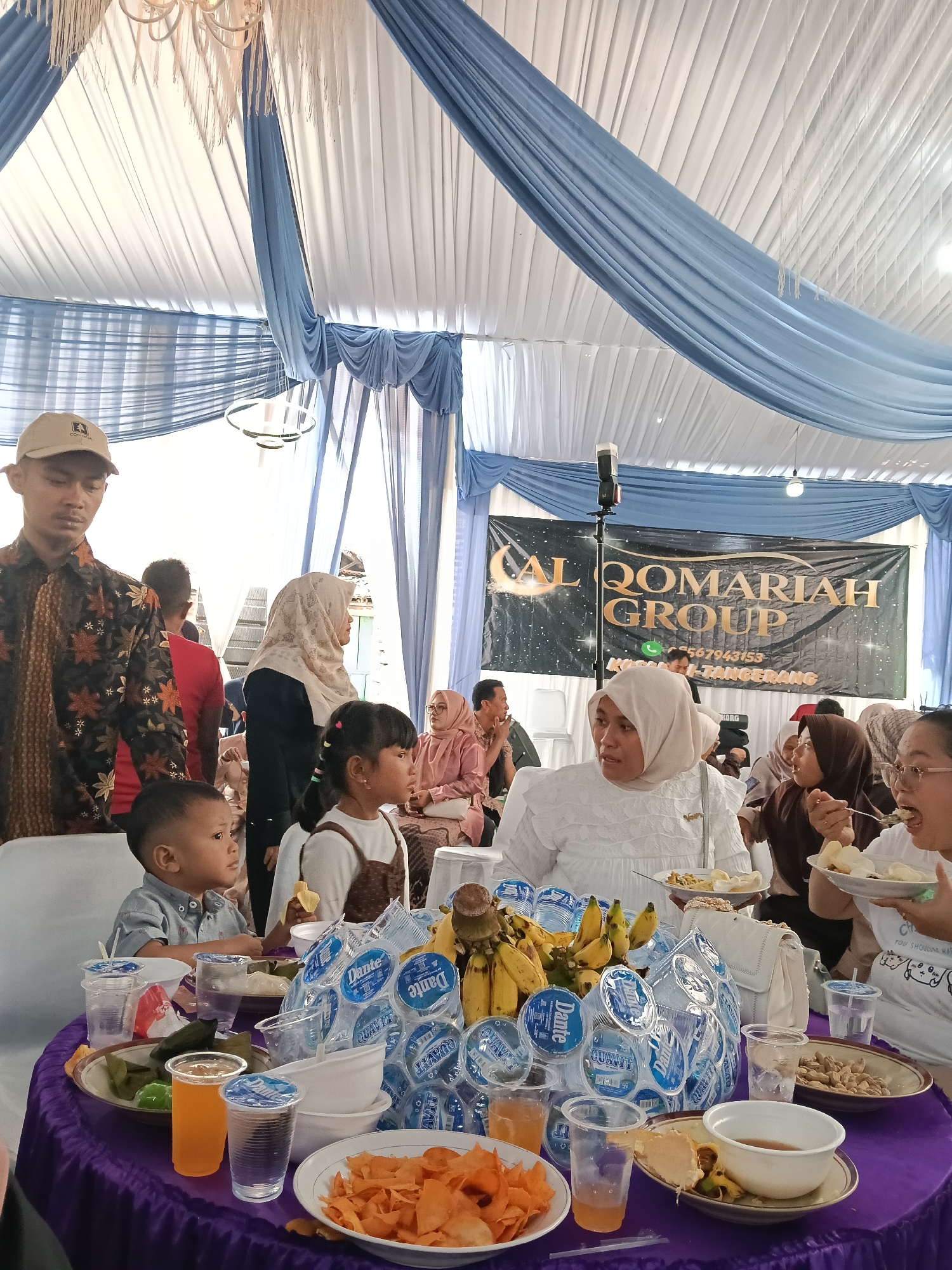 A group of people are seated around a purple-draped round table, attending an event, possibly a wedding reception, under a white and blue draped tent. In the foreground, a young boy in a light blue shirt is seated, looking to his right, with a plate of food and drinks in front of him. A young girl to his right, wearing a brown overall, is also seated and looking to her right. On the table, there are plates of food including what appears to be crackers, bananas, and other dishes, along with several bottles of water and cups with drinks.
Behind the children, several adults are visible. A woman in a white hijab is seated, facing left, with a plate of food. To her right, another woman in a brown hijab is eating with a fork. Further back, a man wearing a patterned batik shirt and a cap stands on the left, looking down. A banner in the background reads "AL QOMARIAH GROUP" with a phone number and "TEGAL ANGUS, TANGERANG". The overall environment suggests an outdoor event in a public space.