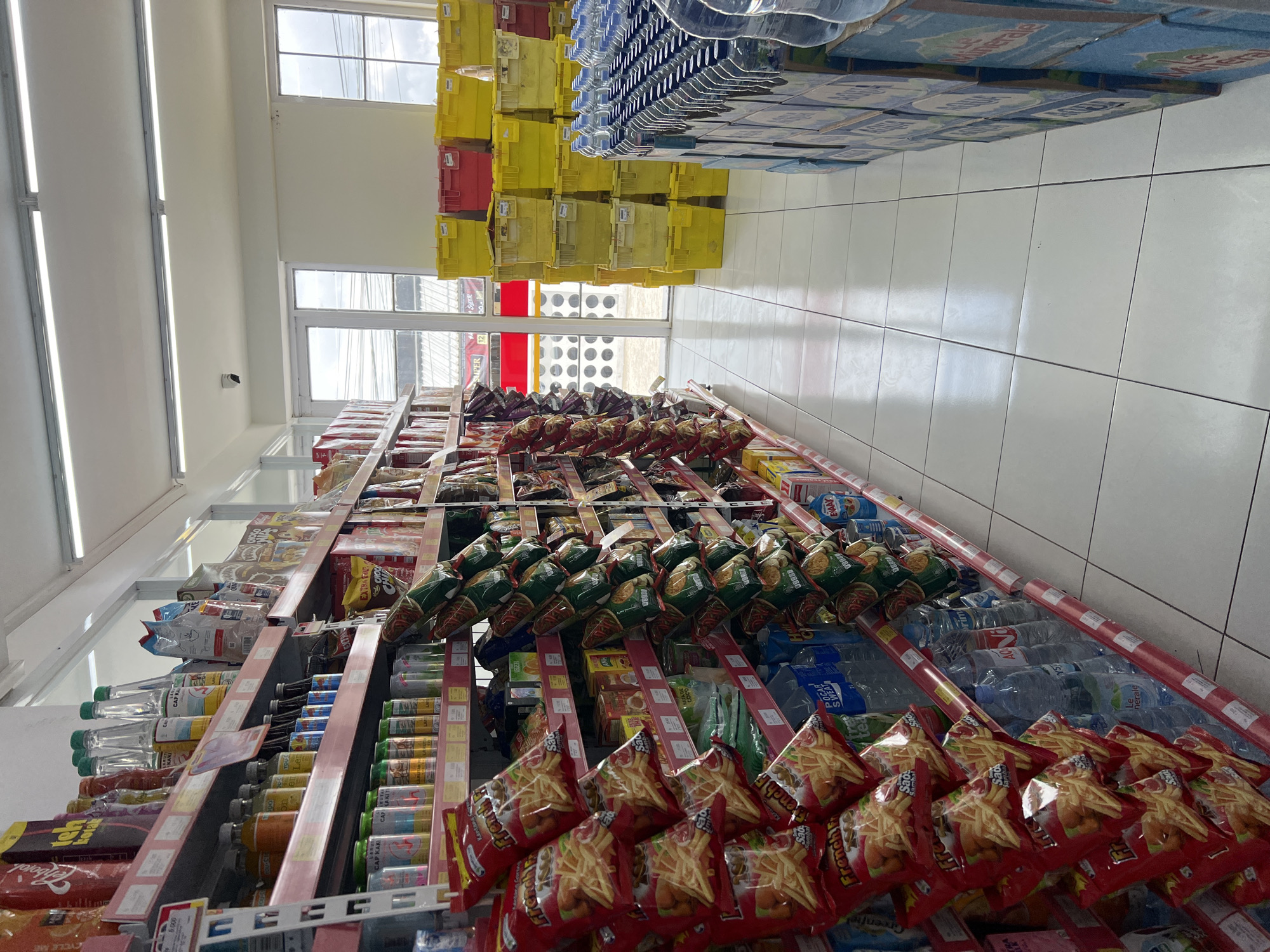 The image depicts the interior of a convenience store aisle in Kintamani, Indonesia. The aisle is lined with shelves stocked with various food and beverage products. On the left, shelves display bags of potato chips, bottles of sauces and drinks, and other packaged goods. In the center and right, more shelves hold similar items, including bottles of water stacked on the right. In the background, through an open doorway, yellow and red plastic crates are visible, along with two windows and a security camera mounted on the ceiling. The floor is tiled in white. No people are present in the scene.
