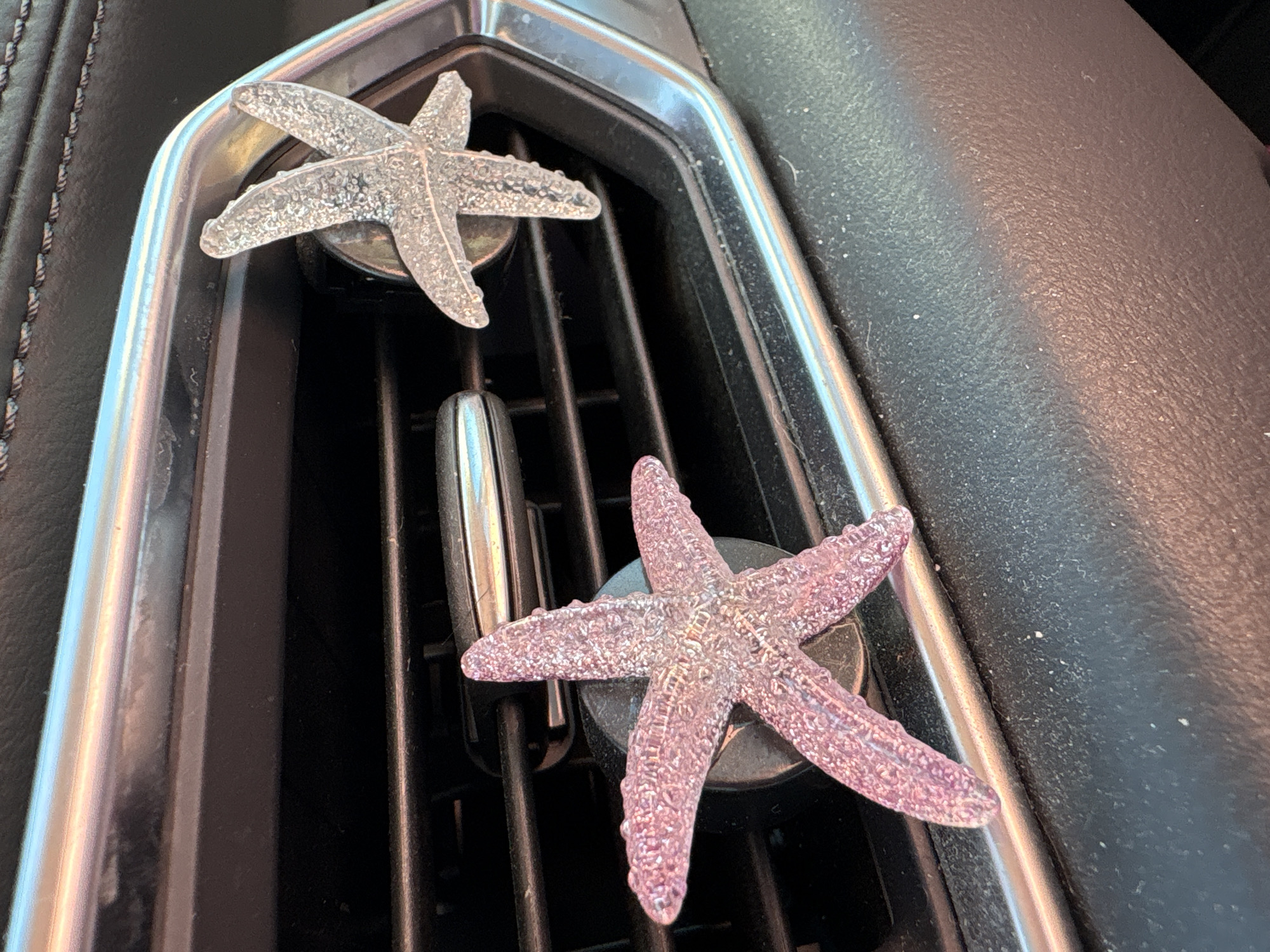 The image shows the dashboard air vent of a car. Two decorative starfish are clipped to the vent. The upper starfish is translucent and colorless. The lower starfish is translucent and light pink. The car’s dashboard is visible on the right side of the image, and a stitched leather section can be seen on the left. This is a generic scene that could be found in any car, and no definitive indicators place this scene specifically in Huntington Beach, California.