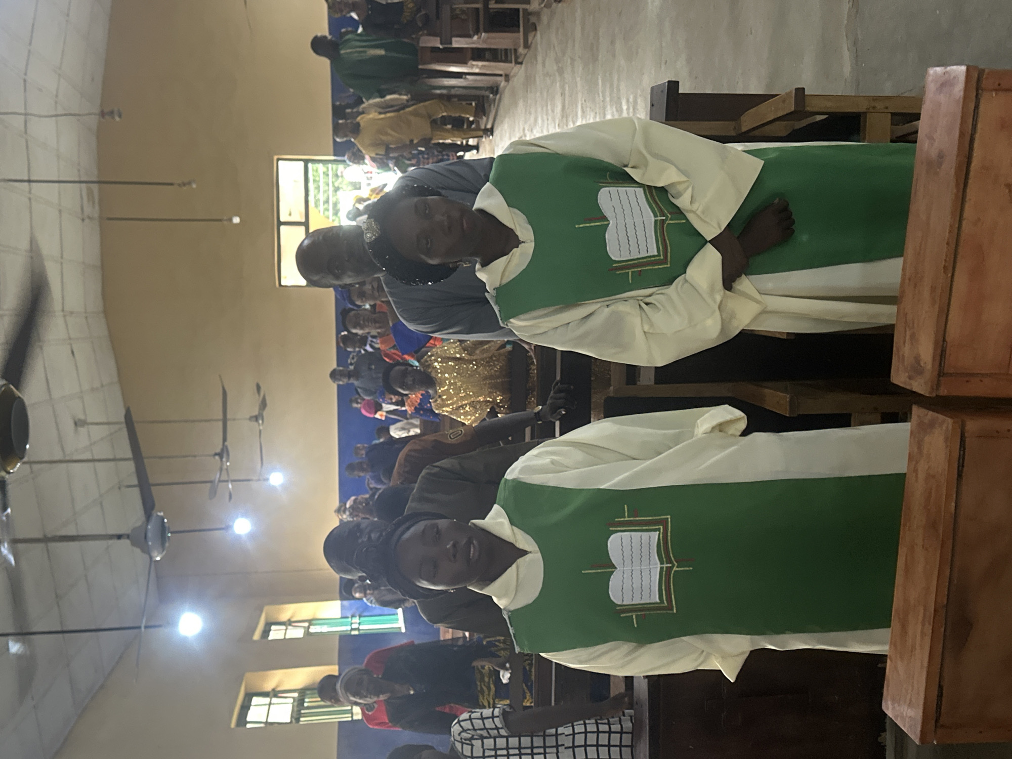 This is a medium shot of two women in white robes and green stoles with an open book emblem, standing in a church in Akwanga, Nigeria. The woman in the foreground on the left has her head slightly bowed and her mouth open as if speaking. The woman on the right is looking forward with a neutral expression. Behind them, several people are standing and some are seated on wooden pews. The church has a tiled ceiling with ceiling fans and fluorescent lights. The walls are painted in a light color, and there are windows with bars visible on the left side of the image. The floor is concrete. In front of the women are two wooden boxes or chests. The overall lighting suggests it is daytime.