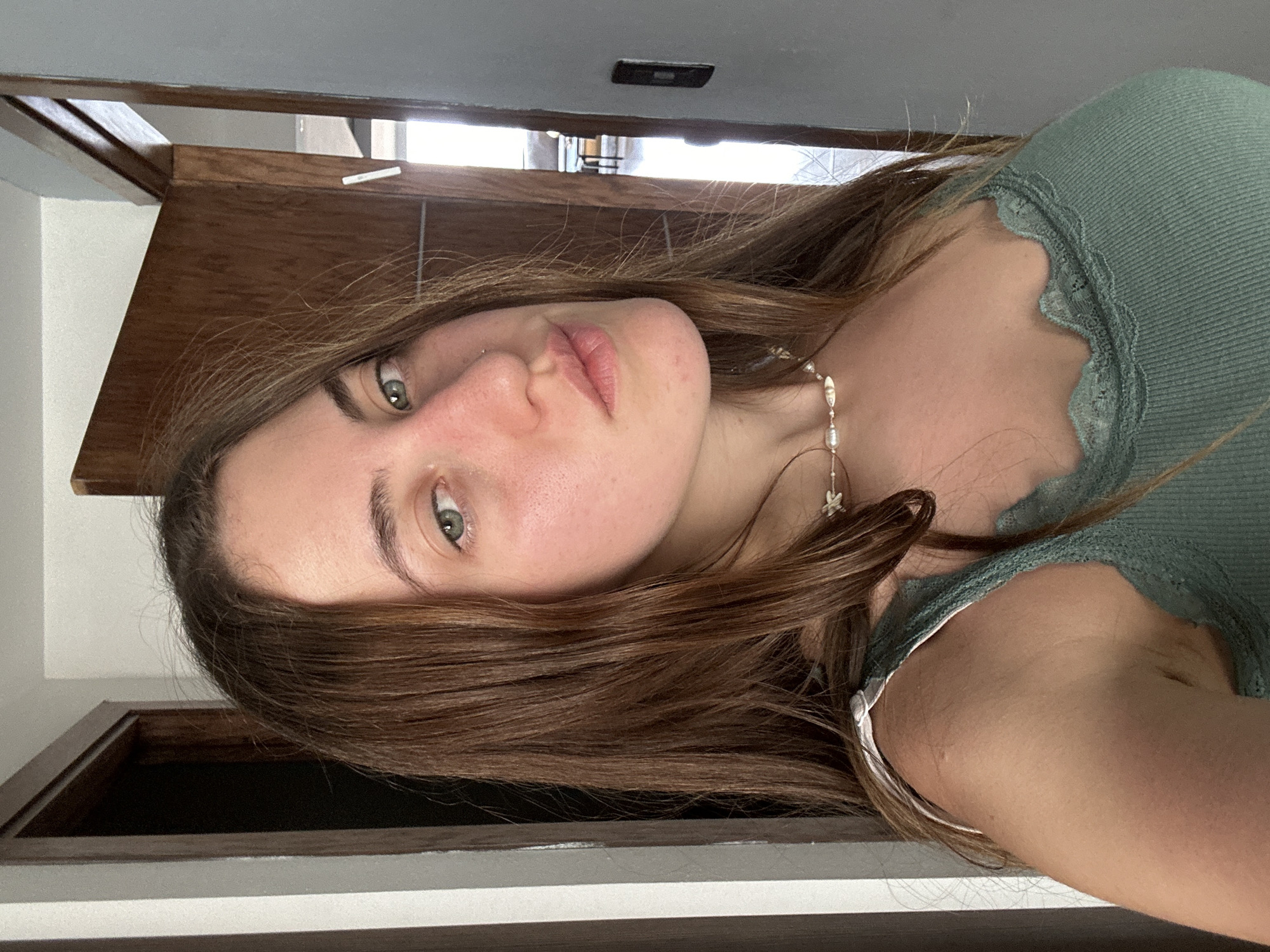 A young woman is pictured indoors. She has brown hair and light green eyes. She is wearing a green tank top with lace detailing around the neckline. She also appears to be wearing a delicate pearl necklace with star charms. The background consists of a wooden door and a grey wall. There is no information to determine the location in the city of Huixquilucan de Degollado, Mexico.