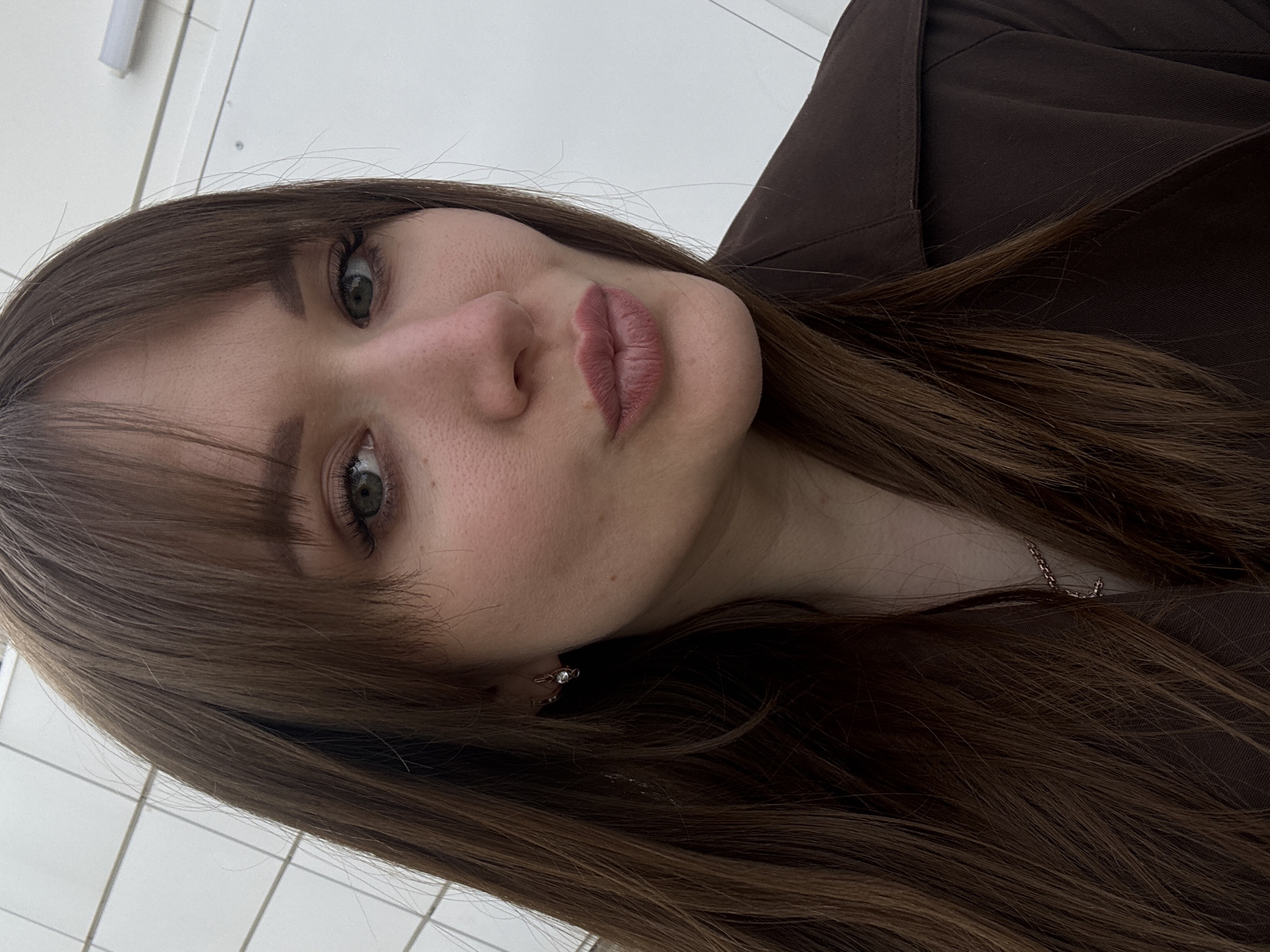A medium close-up shot presents a woman with straight, shoulder-length brown hair and bangs. She has fair skin, green eyes, and a slightly pursed mouth. An earring is visible in her ear. She is wearing a brown garment. A thin silver necklace partially obscured by her hair. The background appears to be a white tiled wall, likely in an interior setting. There is no specific evidence to suggest the photo was taken in Chaikovsky, Russia.