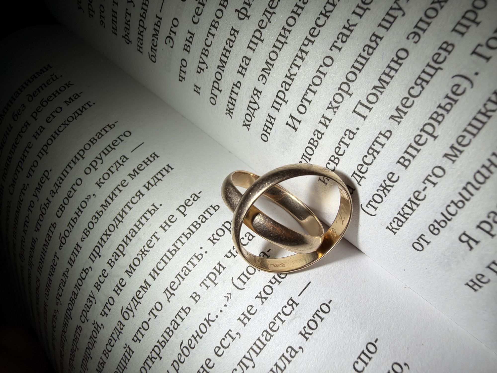 Here is a detailed description of the image:
**Scene Overview:** The image shows an open book with two gold wedding rings resting on the pages. The book appears to be a paperback, and the pages are filled with text. The lighting is subdued, with some areas in shadow, which creates a slightly dramatic effect.
**Subjects/Objects:**
* **Book:** An open book with text written in what appears to be a language other than English, possibly Russian. The pages are slightly worn.
* **Wedding Rings:** Two gold wedding rings are intertwined, resting on the open pages of the book. One ring is slightly above and overlaps the other.
* **Text:** The pages are densely filled with text.
**Actions/Interactions:** The rings are placed on the book. No people are directly visible.
**Environment:** The setting is indistinct, but the presence of a book suggests an interior setting, such as a study, library, or home.
**Overall Impression:** The image has a sentimental or symbolic quality. The combination of wedding rings and text could suggest themes of love, commitment, and shared stories.